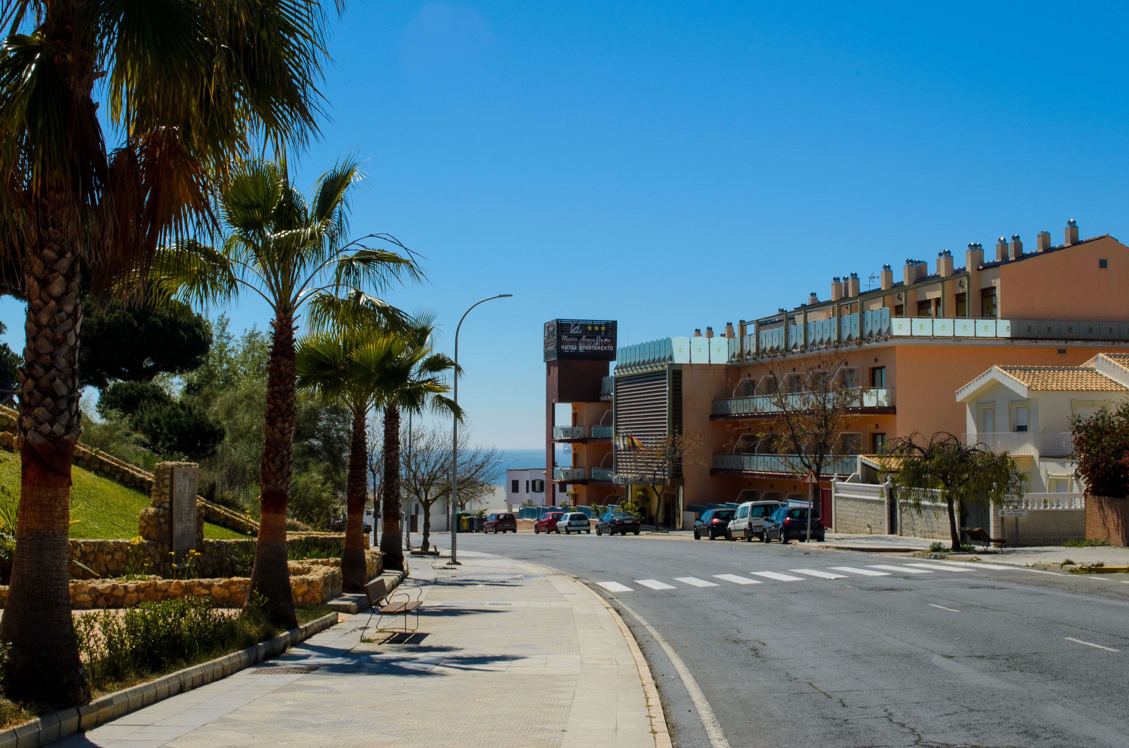 Vista de la calle con un hotel que tiene balcones, cielo despejado y coches estacionados.