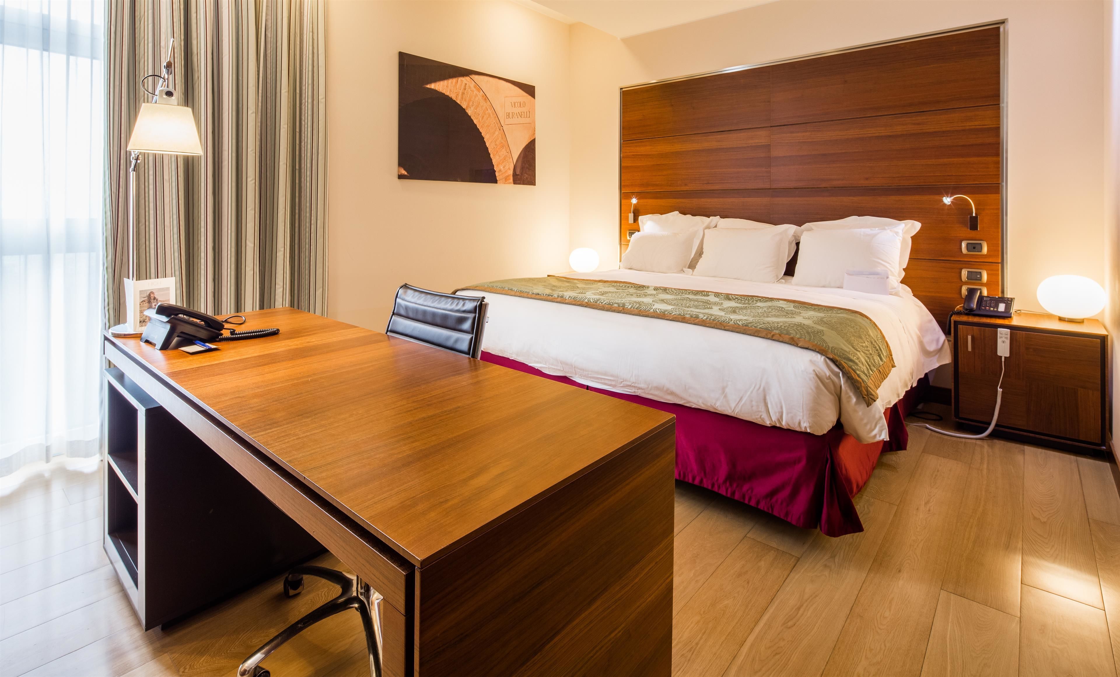 Ein Hotelzimmer mit Holzschreibtisch, einem Bett mit weißer Bettwäsche, einem Nachttisch mit Lampe und einem Gemälde an der Wand.