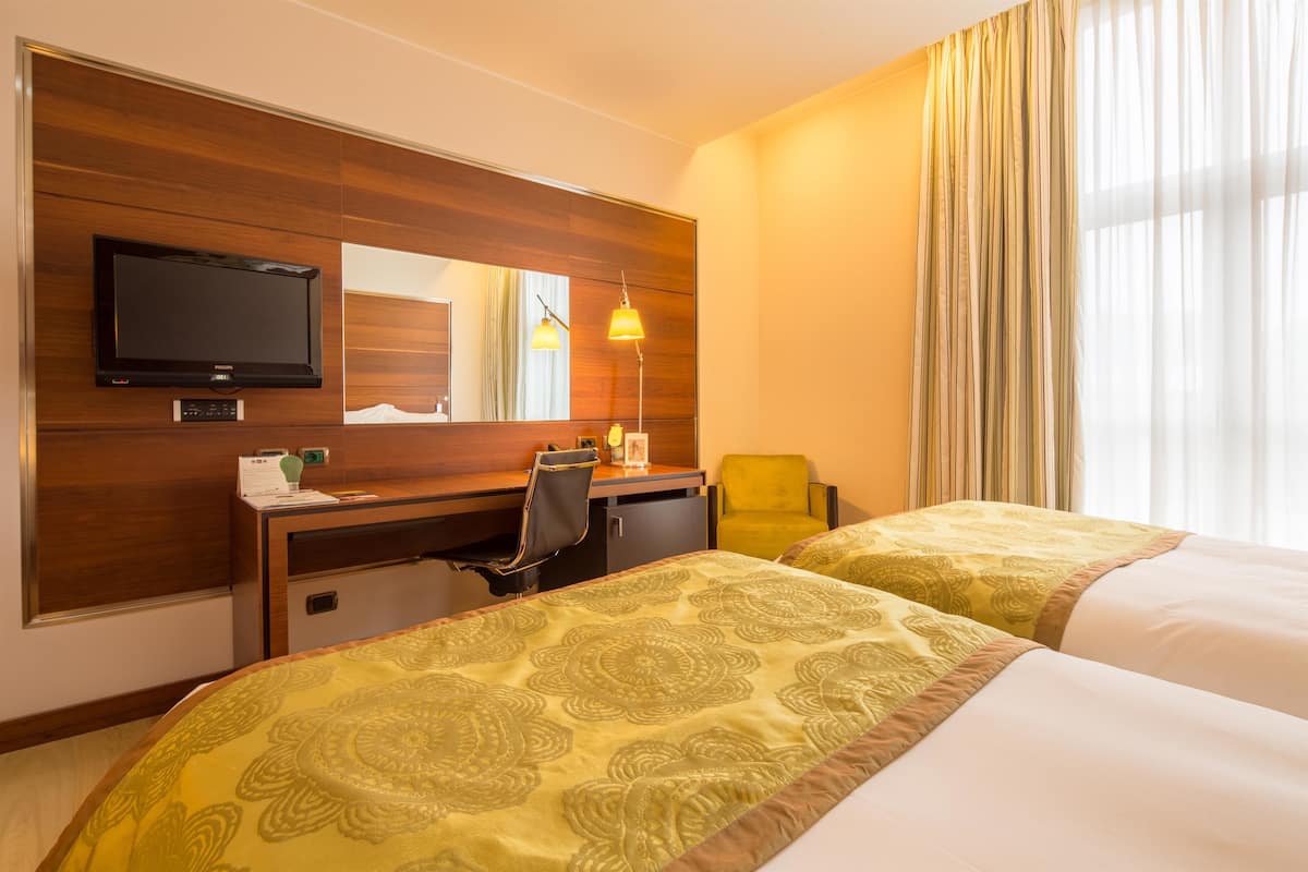 Habitación de hotel con dos camas, un escritorio, una silla, un televisor y un ventanal con cortinas.
