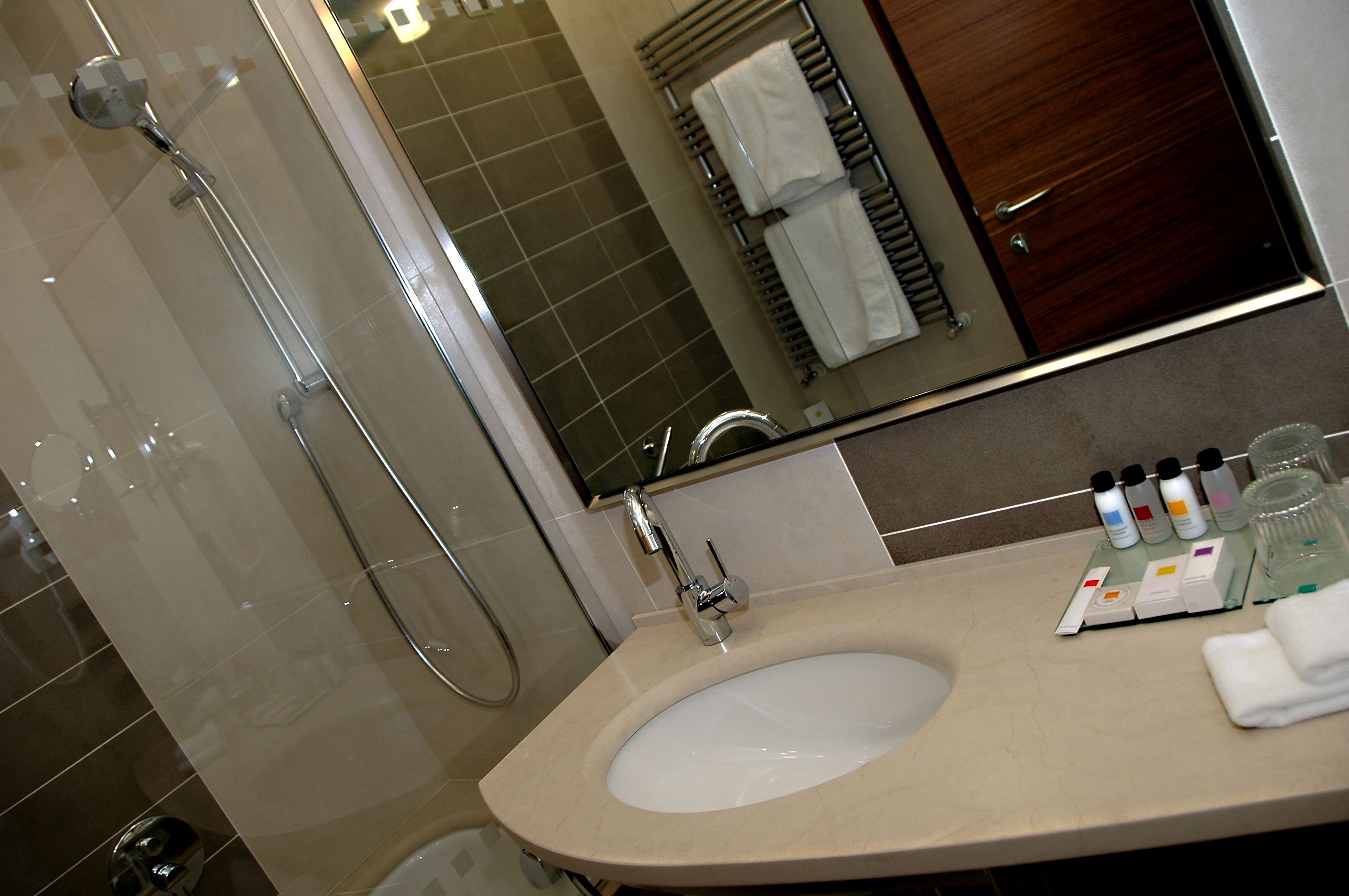 Une salle de bain dotée d’une douche à parois de verre, d’un lavabo avec des articles de toilette et d’un miroir qui reflète un porte-serviettes et une porte en bois.