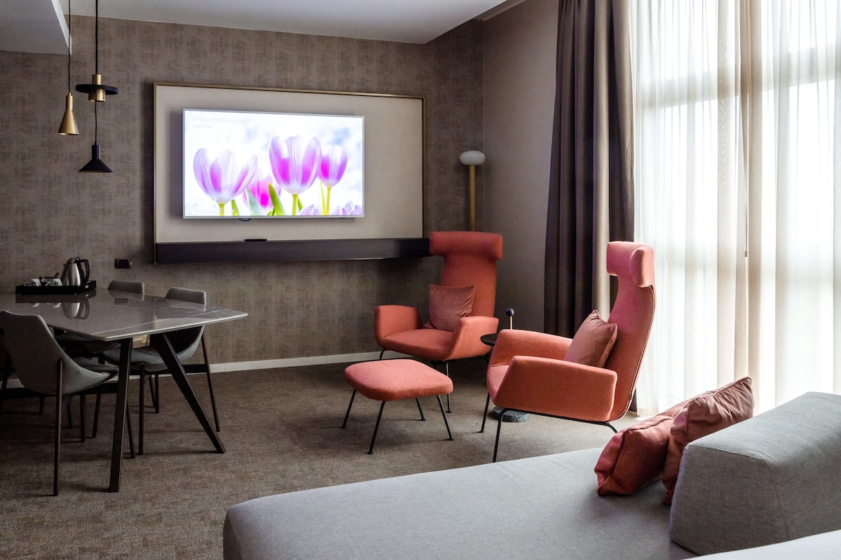 Una habitación de hotel moderna con una televisión de pantalla plana en la pared, una mesa de comedor con sillas, un sofá y un ventanal con cortinas.