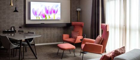 Une chambre d’hôtel moderne dotée d’un téléviseur à écran plat fixé au mur, d’une table à manger avec des chaises, d’un canapé et d’une grande fenêtre avec des rideaux.