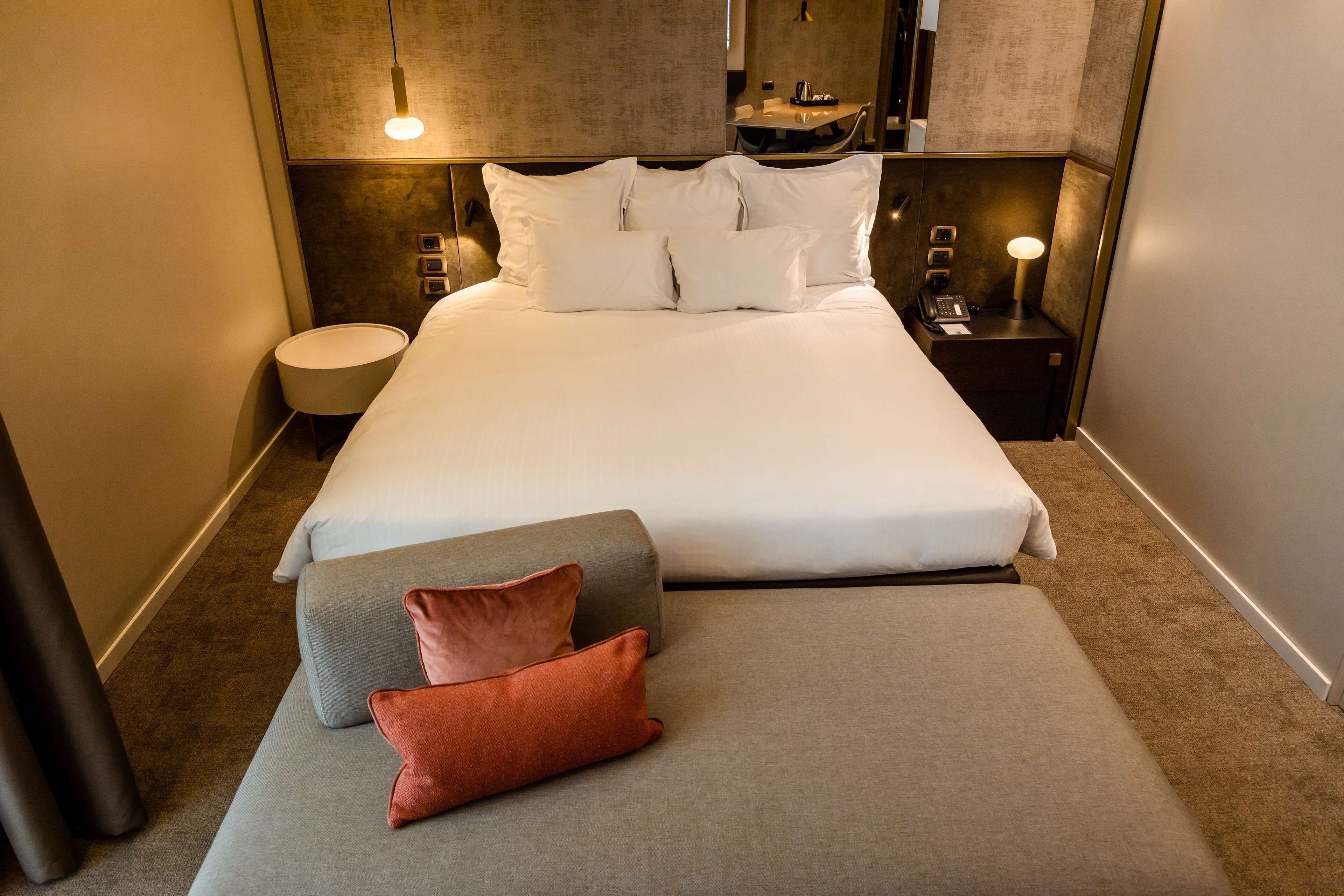 Una cama bien hecha con ropa blanca, una almohada decorativa gris y roja, y una mesita de noche con una lámpara y un teléfono.