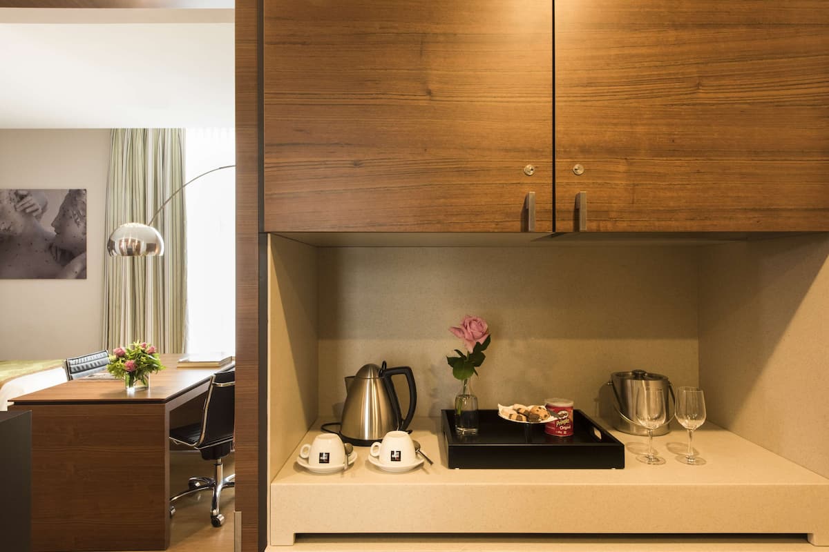 Habitación de hotel con un escritorio de madera, una silla, una cafetera, un florero con una flor, una bandeja con una taza y un vaso, y una copa de vino.