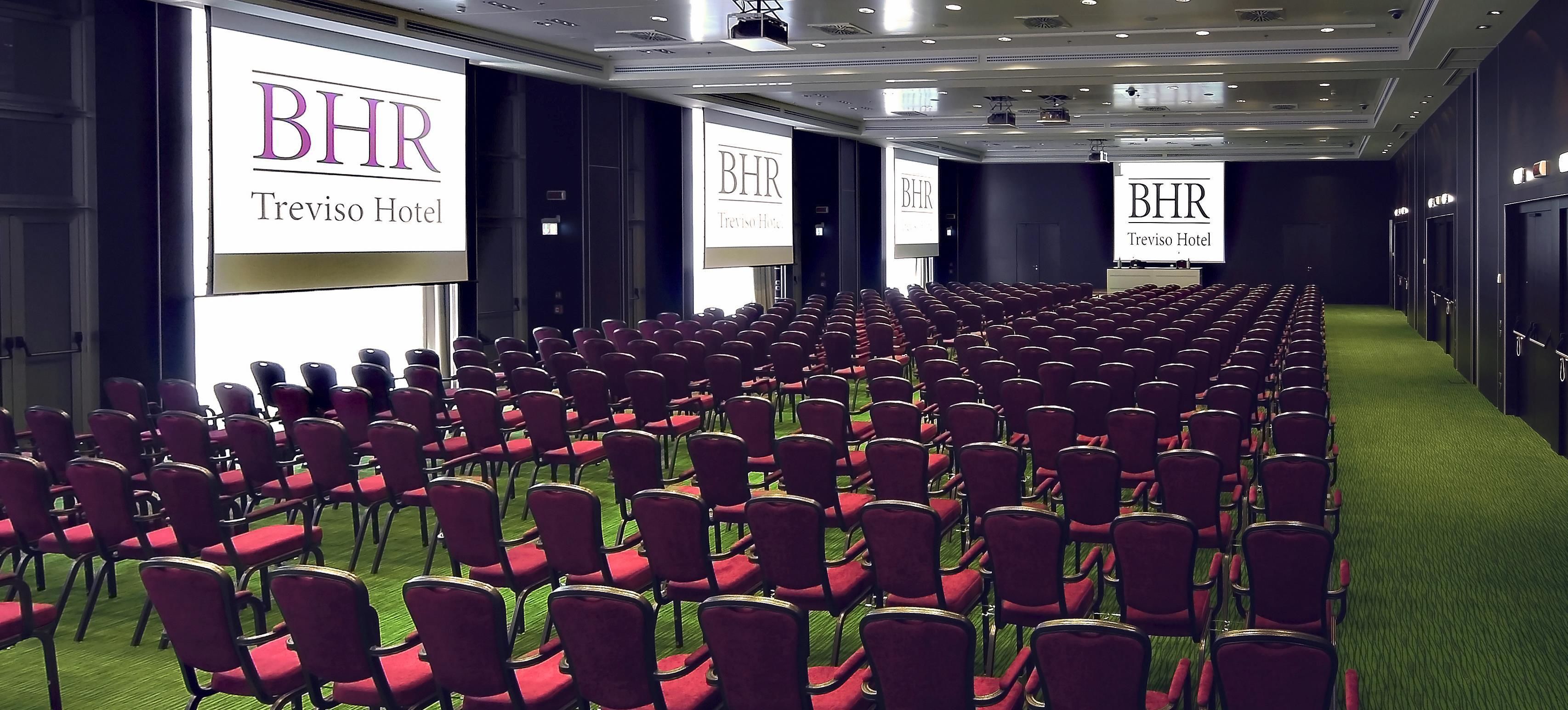 A conference room at Treviso Hotel with rows of red chairs and large screens displaying the hotel’s name.