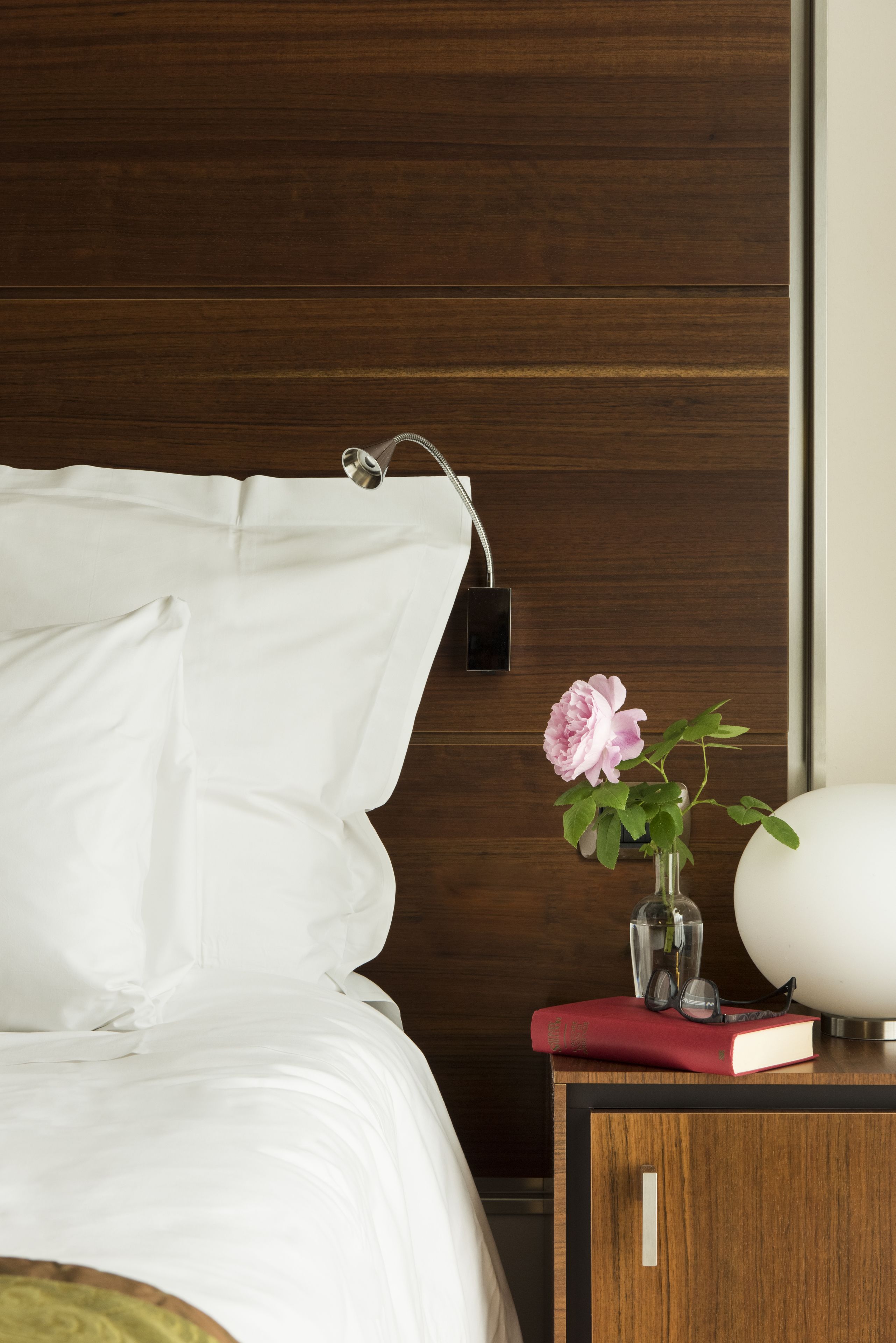 A neatly made bed with white linens, a wooden nightstand with a vase of pink flowers, a book, and a lamp.