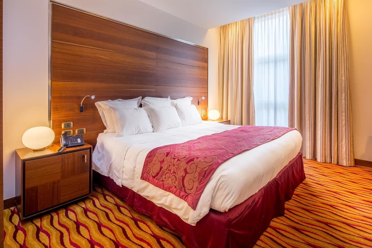 Una habitación de hotel con cabecera de madera, cama grande con ropa de cama blanca y roja, dos lámparas de noche, una mesita de noche y alfombra a rayas.