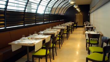 Un restaurante con mesas largas, sillas acolchadas de color verde y un techo curvo con tragaluces.