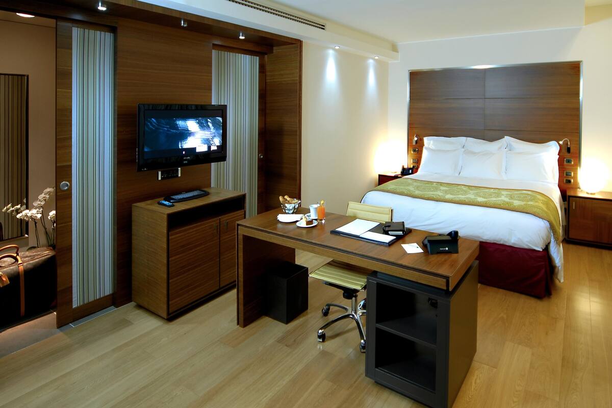 Habitación de hotel con cama, escritorio con computadora, televisión y un pequeño rincón de descanso con una silla y una mesa.