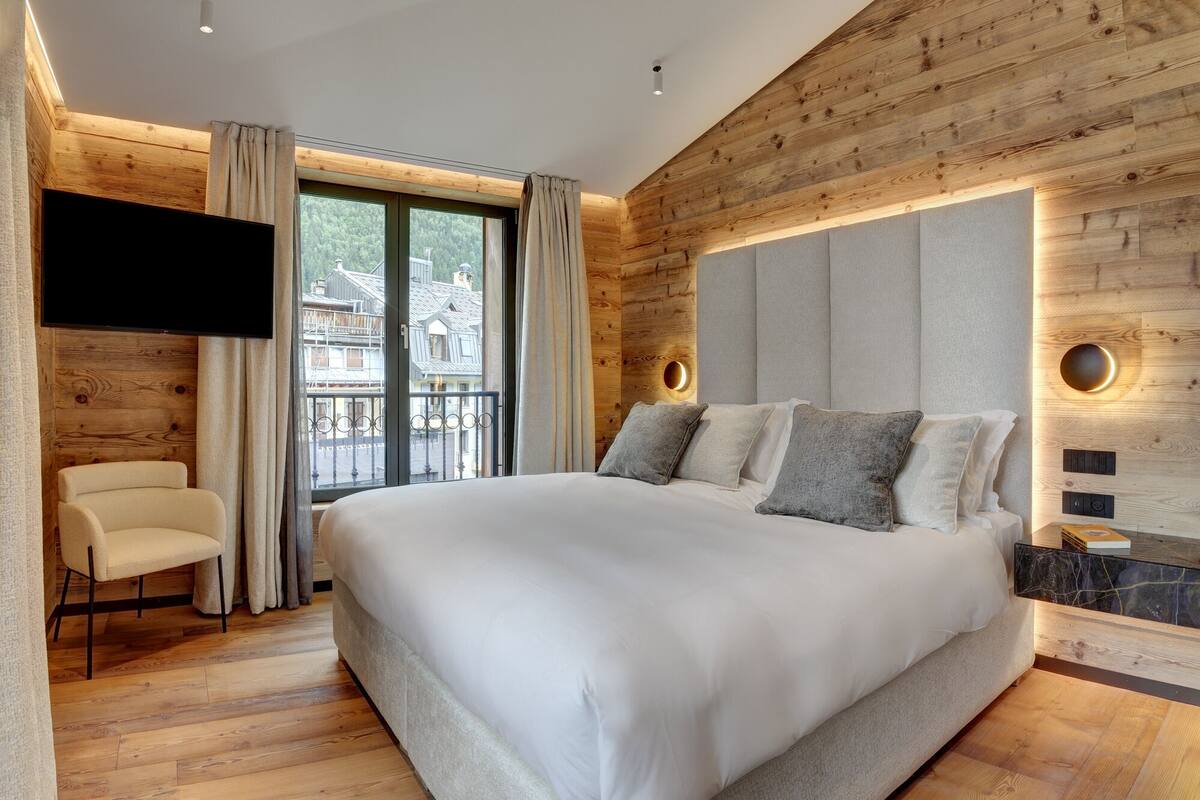 Ett modernt sovrum med en stor säng, en tv som är monterad på väggen och utsikt över en balkong med omgivande byggnader.