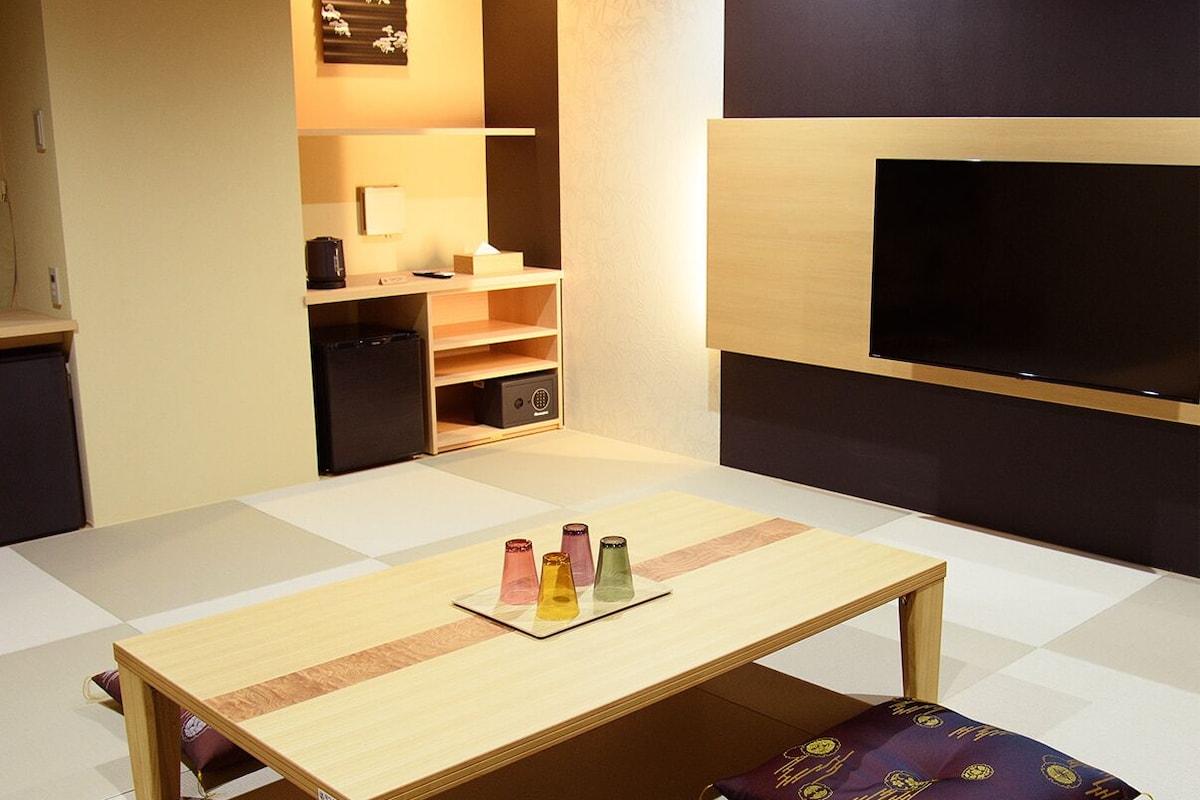 A compact room with a wooden table, a sofa, a TV, and a small kitchenette.
