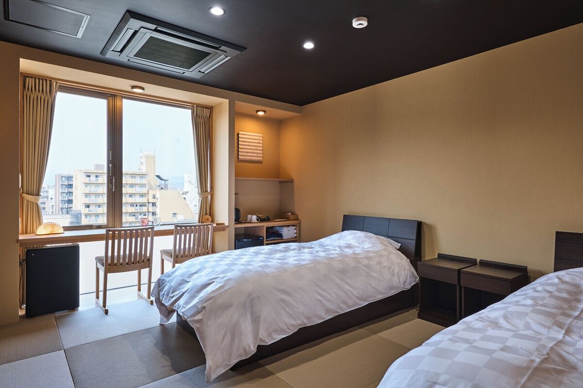 A hotel room with a bed, a desk, a chair, a window with curtains, and a view of buildings.