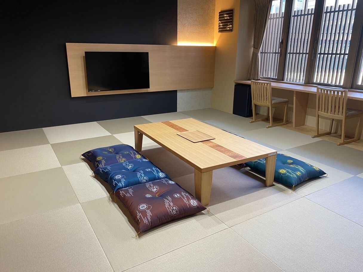 Um espaço com esteiras de tatame, uma mesa baixa, almofadas, uma TV fixada na parede e uma janela com persianas.