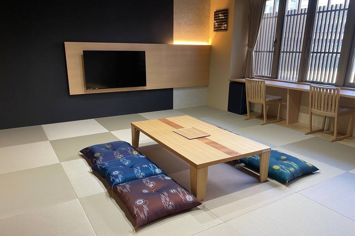 A tatami mat room with a low table, cushions, a mounted TV, and a window with blinds.