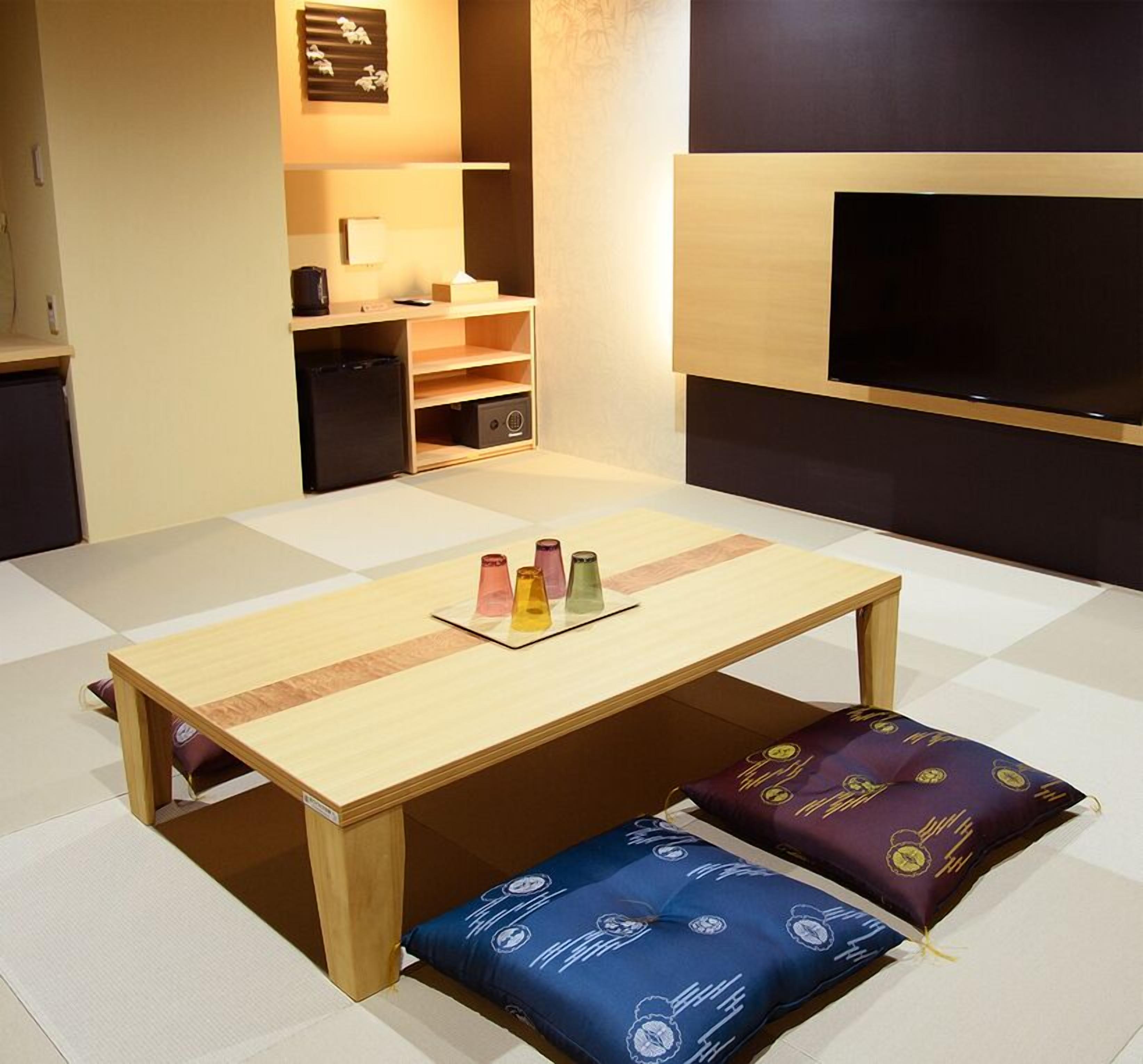 Quarto compacto com mesa de madeira, almofadas coloridas, pequeno espaço de cozinha e televisão de tela plana.