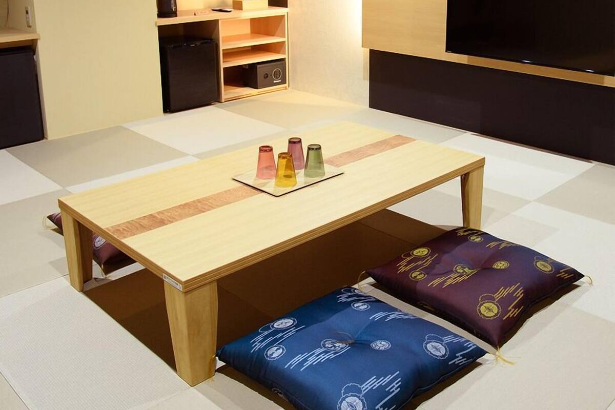A compact room with a wooden table, colorful cushions, a small kitchenette, and a flat-screen TV.