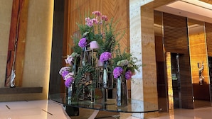 Un lobby moderno con una mesa de vidrio central decorada con arreglos florales, paneles de madera en las paredes y un piso de mármol pulido.
