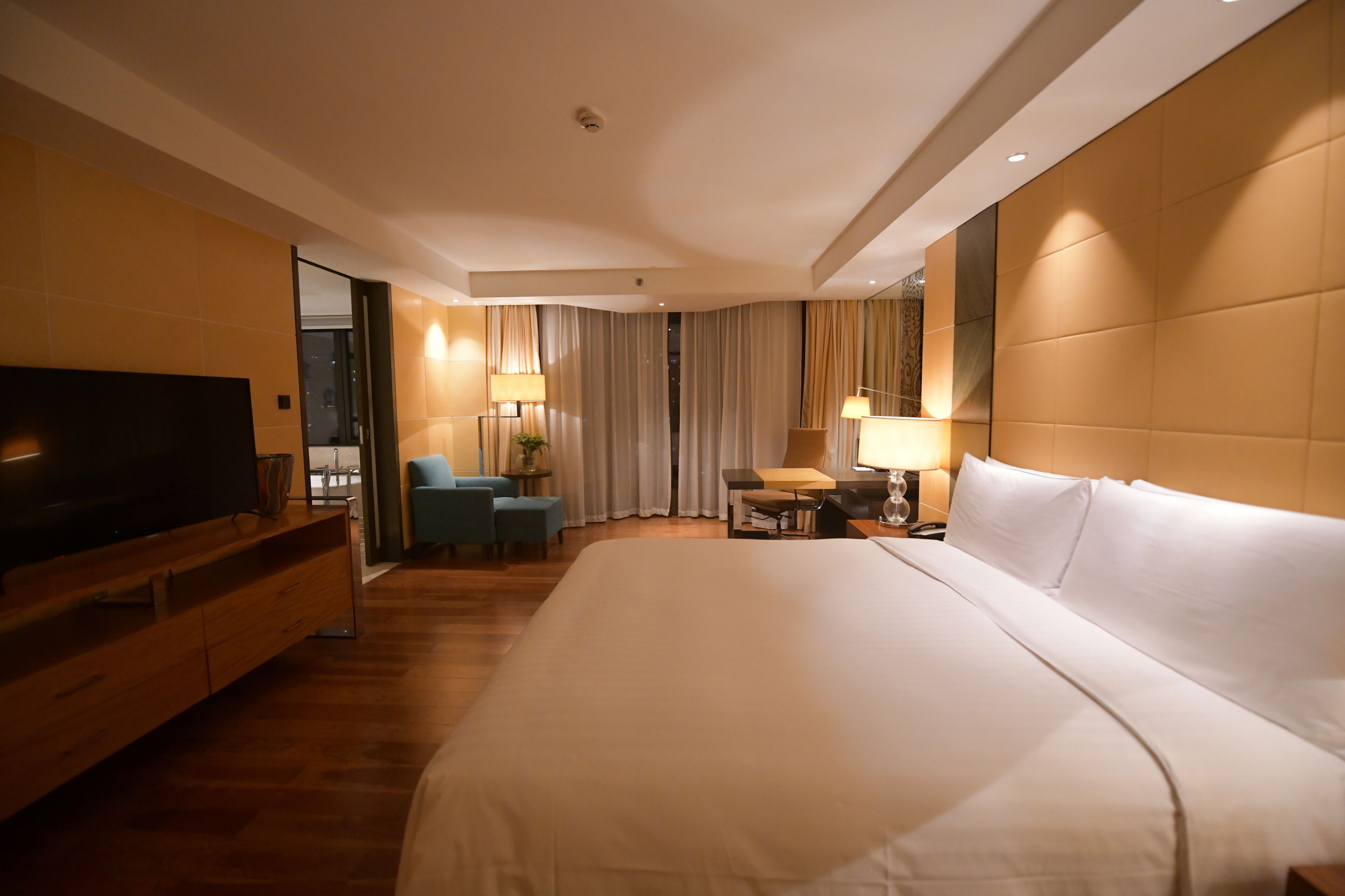 A hotel room with a large bed, a flat-screen TV on a wooden dresser, a desk with a chair, and a window with curtains.