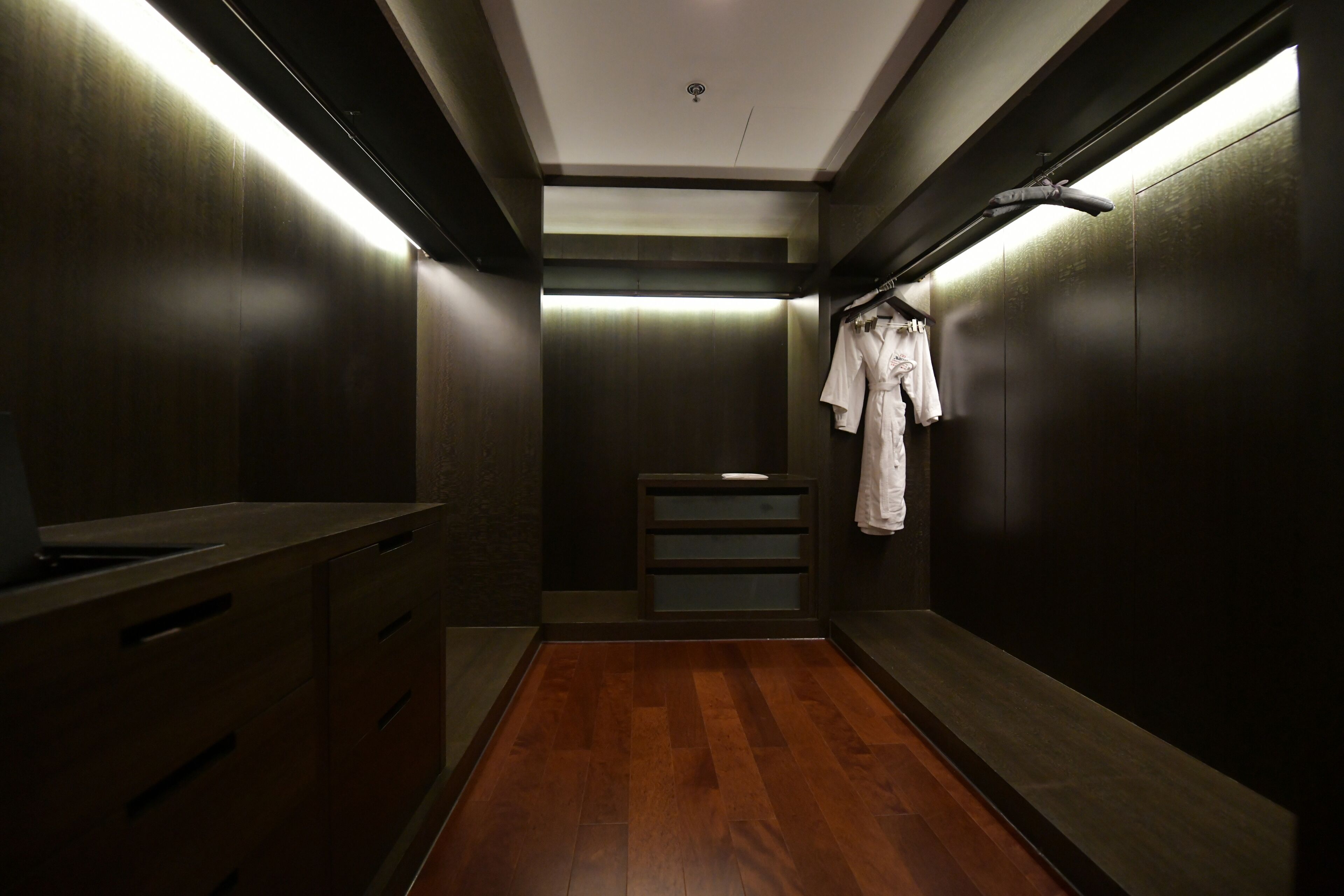 A narrow, dimly lit hallway with wooden flooring and walls, leading to a small seating area with a bench.