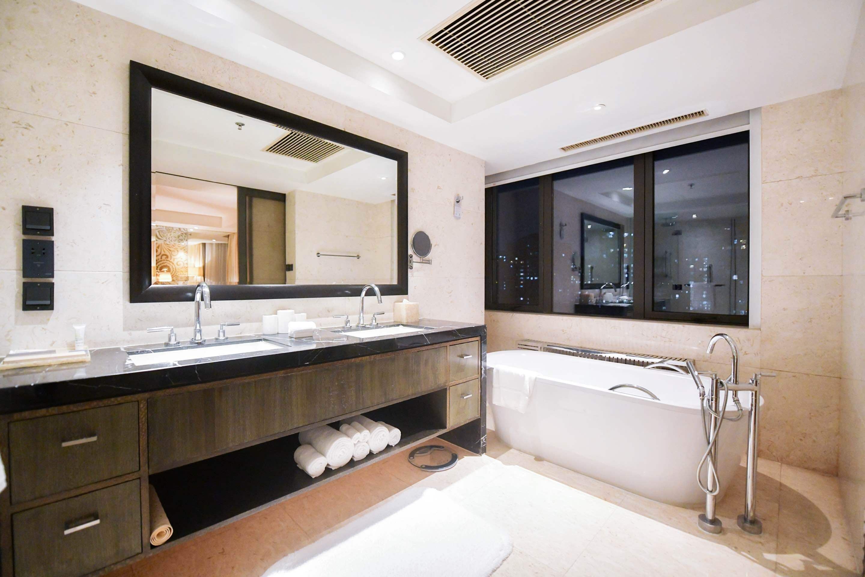 A modern bathroom with a large mirror, dual sinks, a bathtub, and a window with a city view.