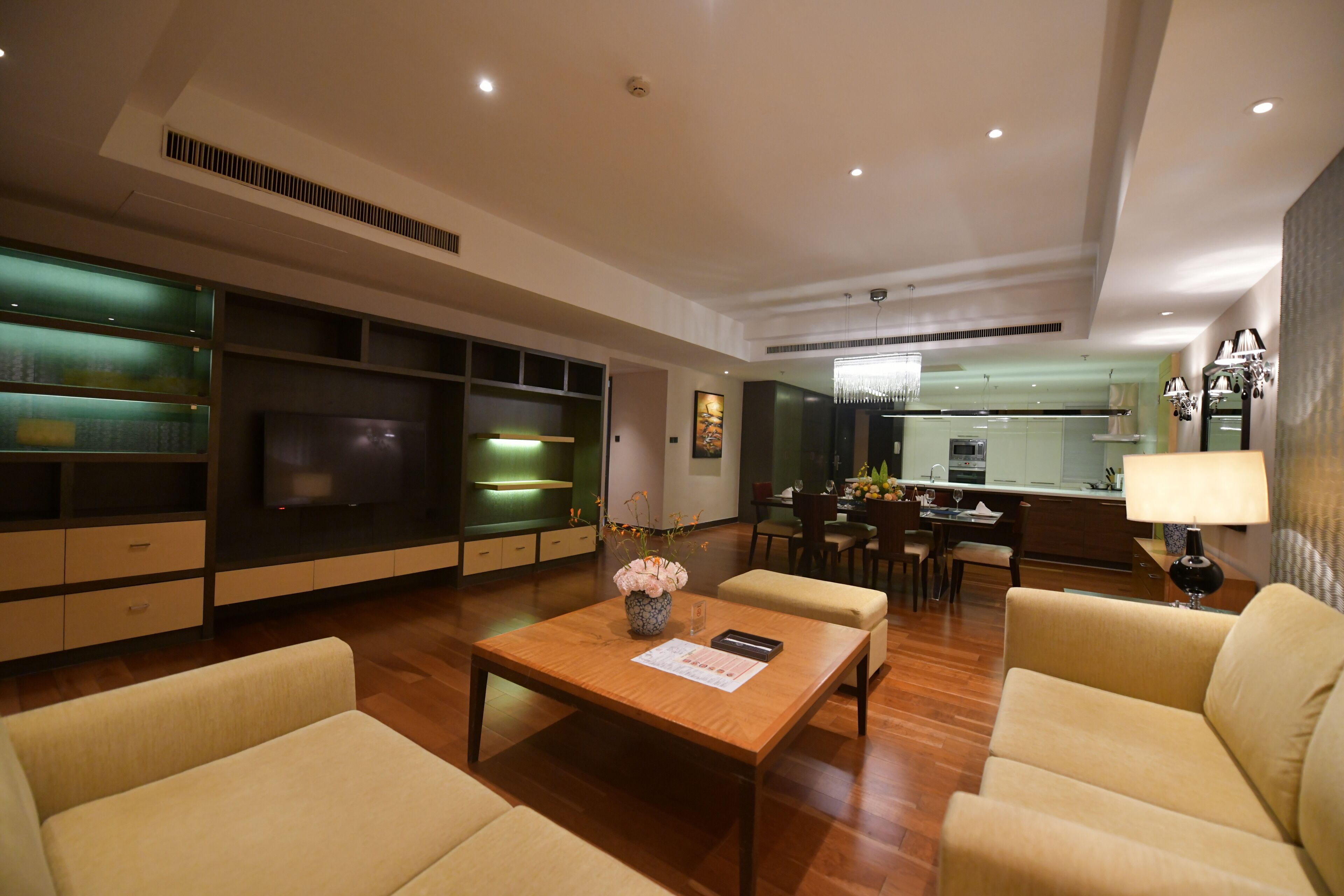A modern living room with a wooden coffee table, beige sofas, a flat-screen TV, and a kitchen area in the background.