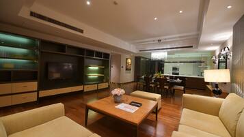 A modern living room with a wooden coffee table, beige sofas, a flat-screen TV, and a kitchen area in the background.
