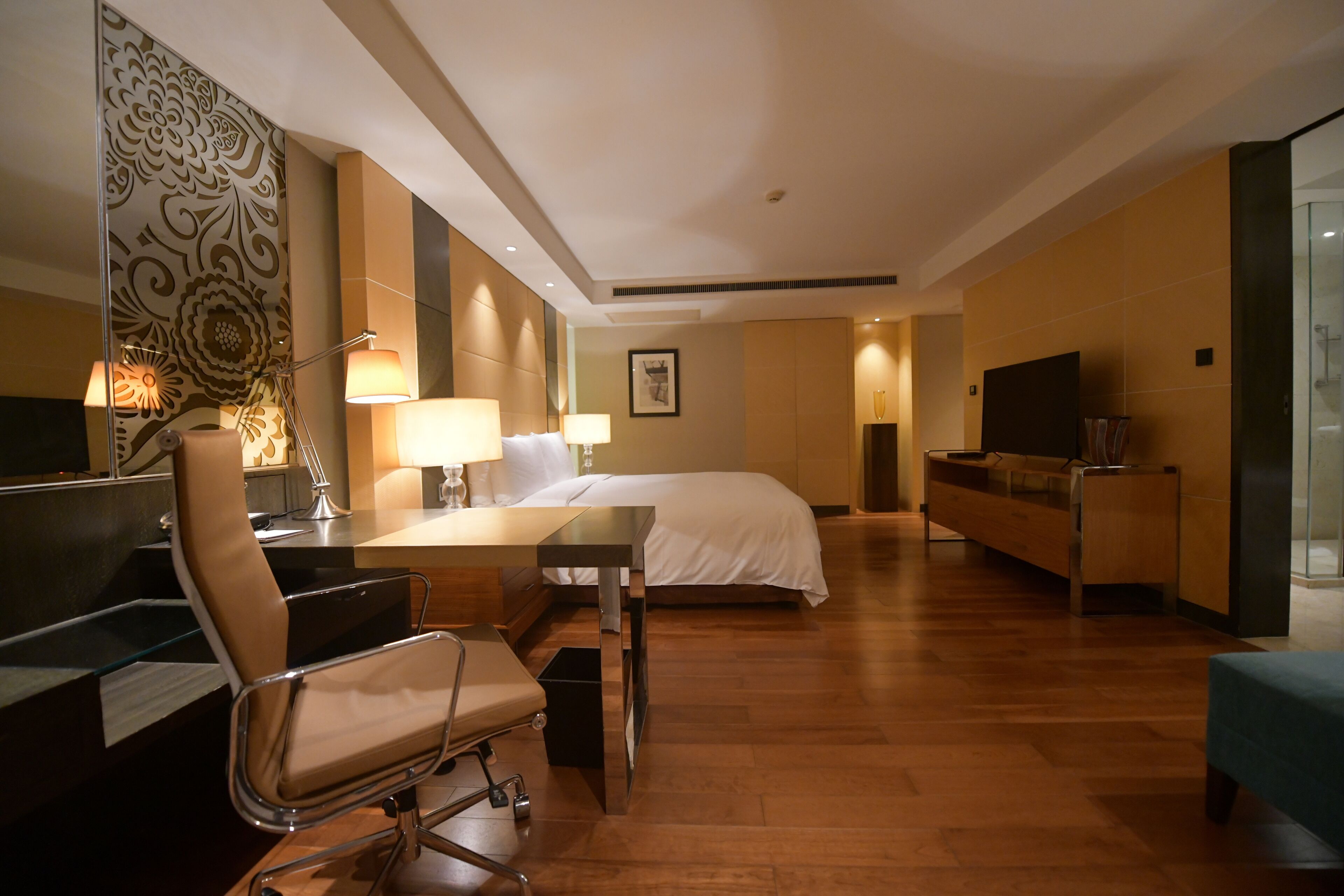 A hotel room with a bed, desk, chair, TV, and a glass shower area.