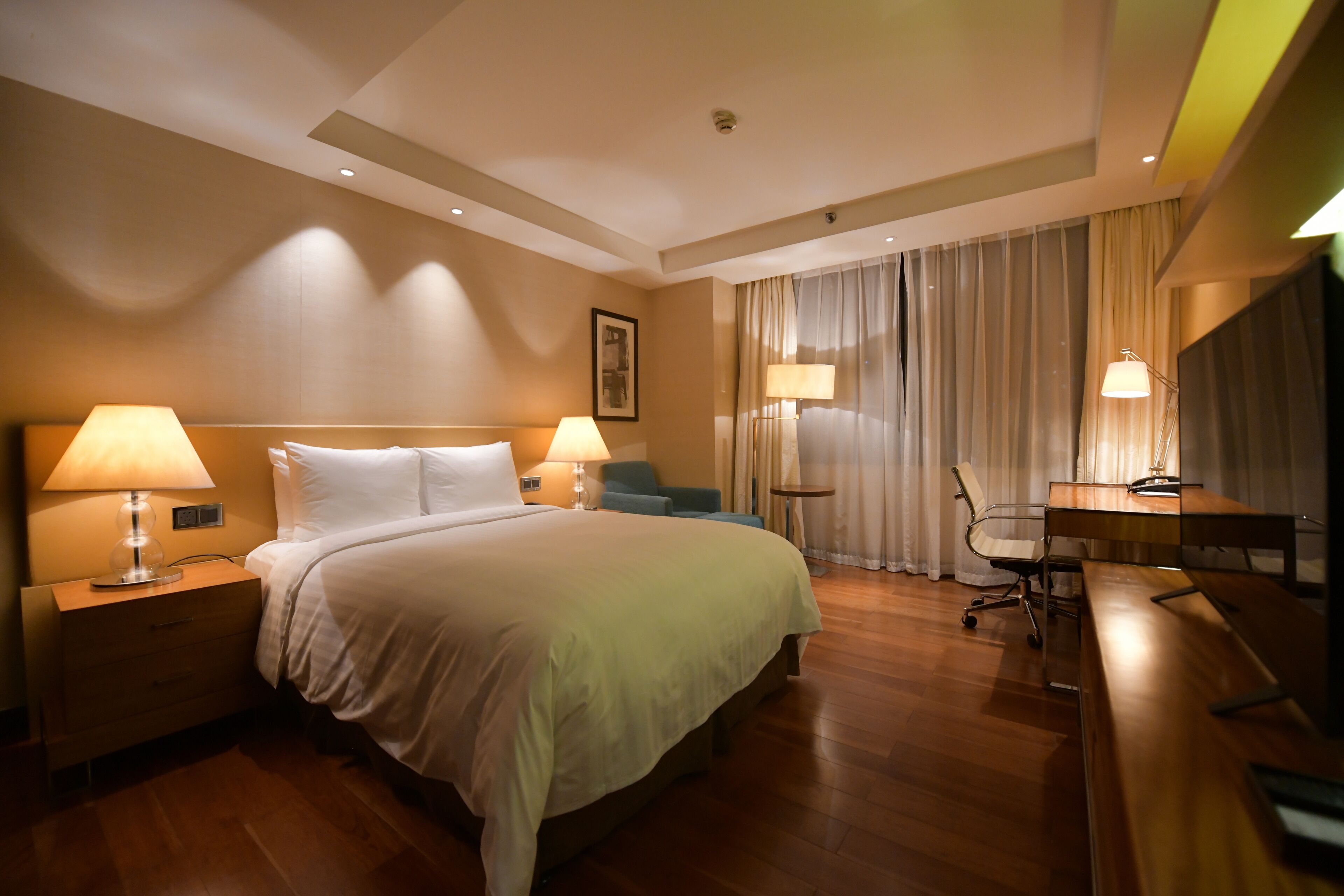 Quarto de hotel com uma cama grande, duas mesas de cabeceira com luminárias, uma escrivaninha com cadeira e uma janela ampla com cortinas.