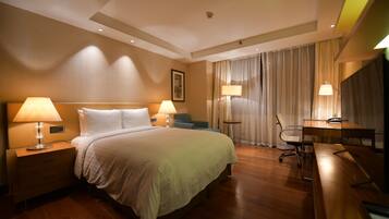 Quarto de hotel com uma cama grande, duas mesas de cabeceira com luminárias, uma escrivaninha com cadeira e uma janela ampla com cortinas.