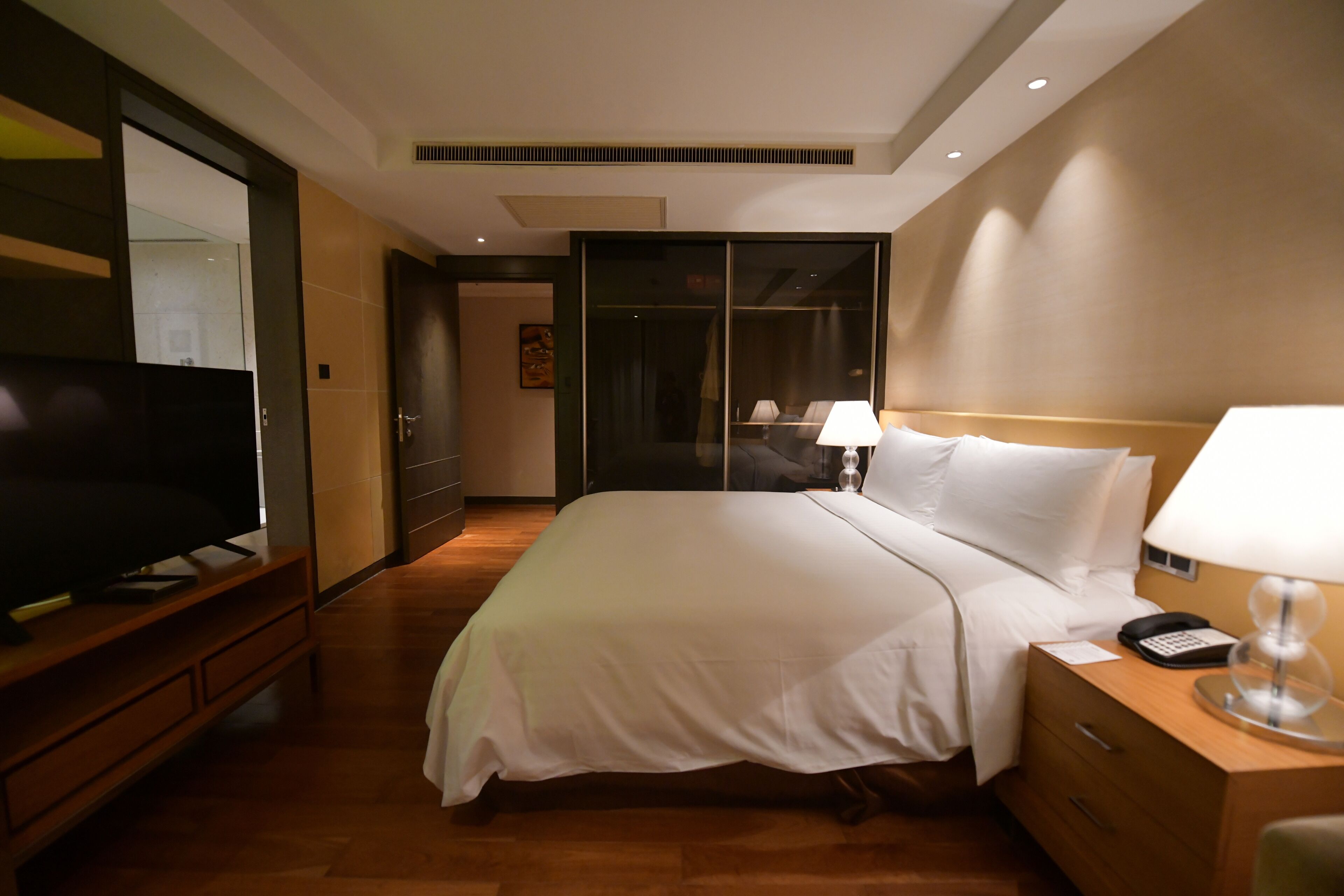Una habitación de hotel con una cama grande, lámparas de noche, un televisor sobre un soporte de madera y un armario con espejo.