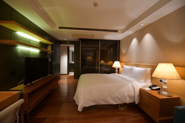 Habitación de hotel con cama, lámparas de noche, televisor sobre un soporte de madera y mesita de noche de madera.