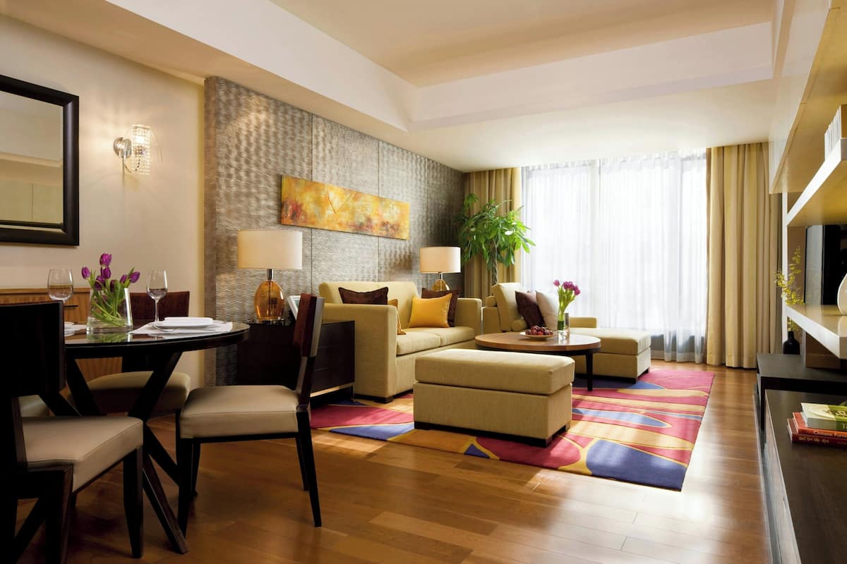 A modern hotel room with a dining area, a sofa set, a television, and a large window with curtains.
