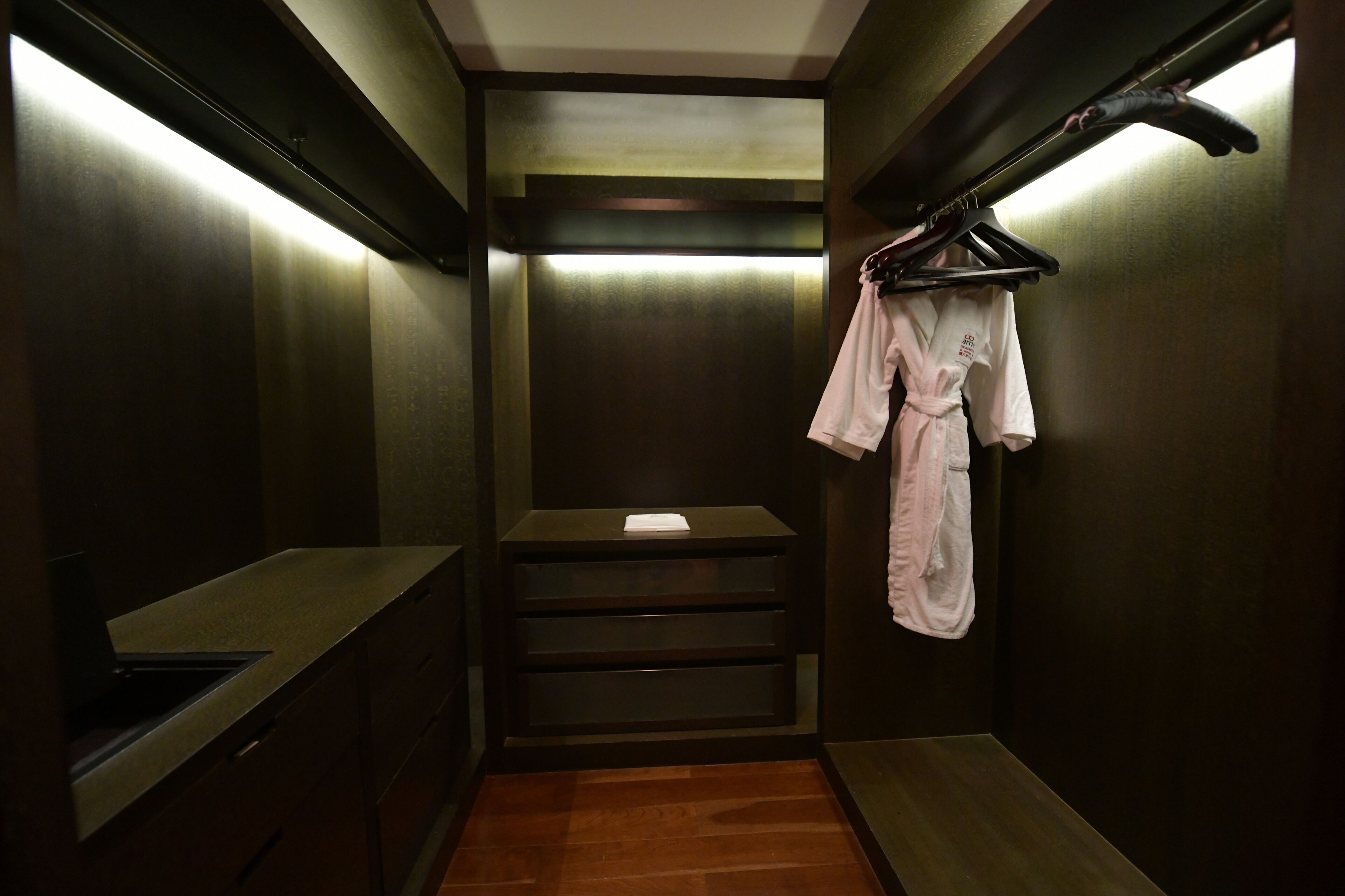 A narrow, dimly lit hallway with a bench, a small table with a piece of paper, and a clothing rack with a robe.