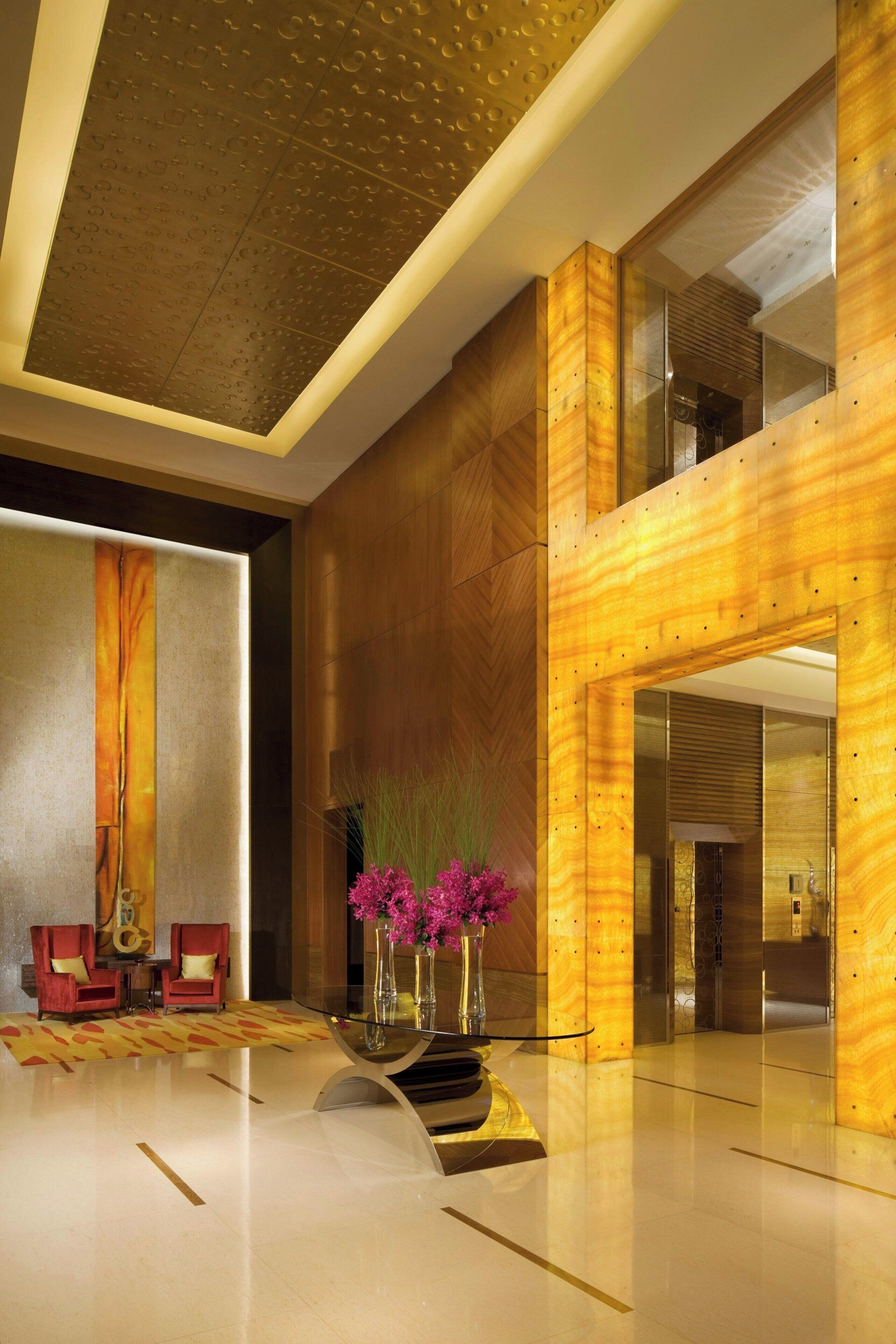 A modern lobby with a wooden reception desk, two red armchairs, a glass coffee table, and a vase with pink flowers.