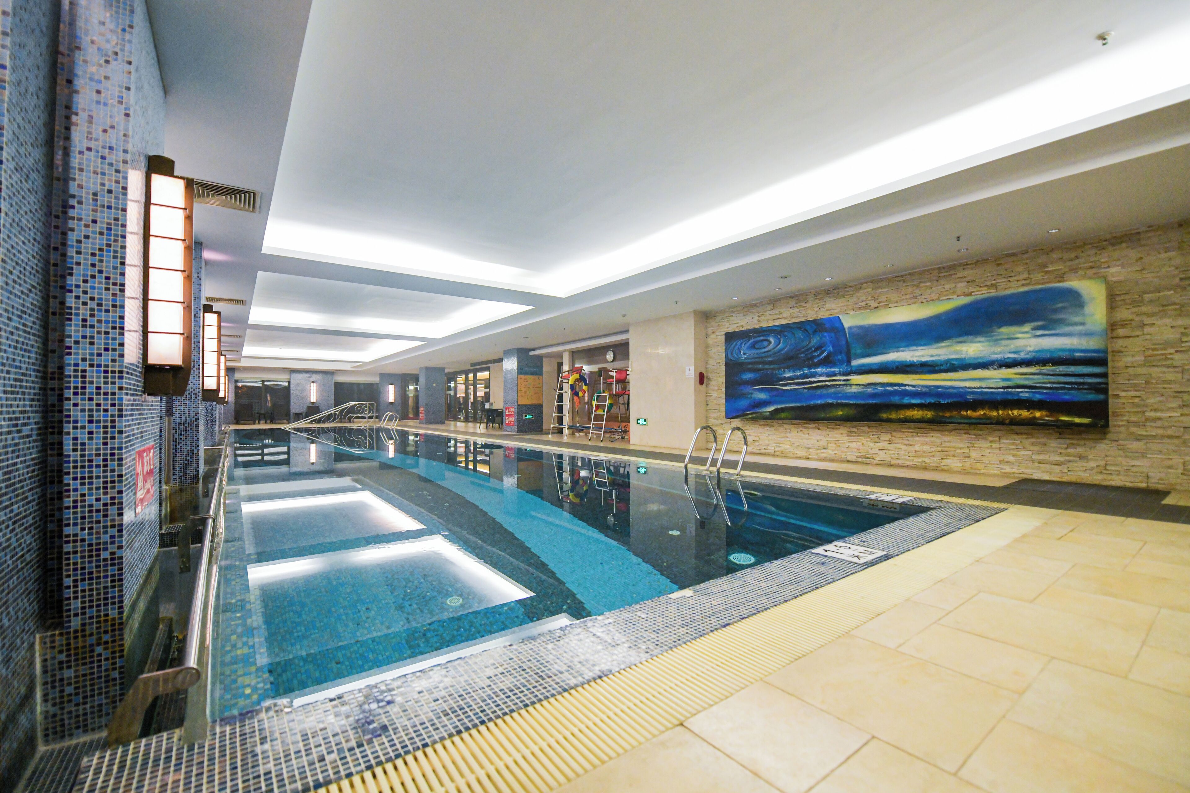 Piscina interior com água cristalina, paredes azulejadas e um grande mural à direita.