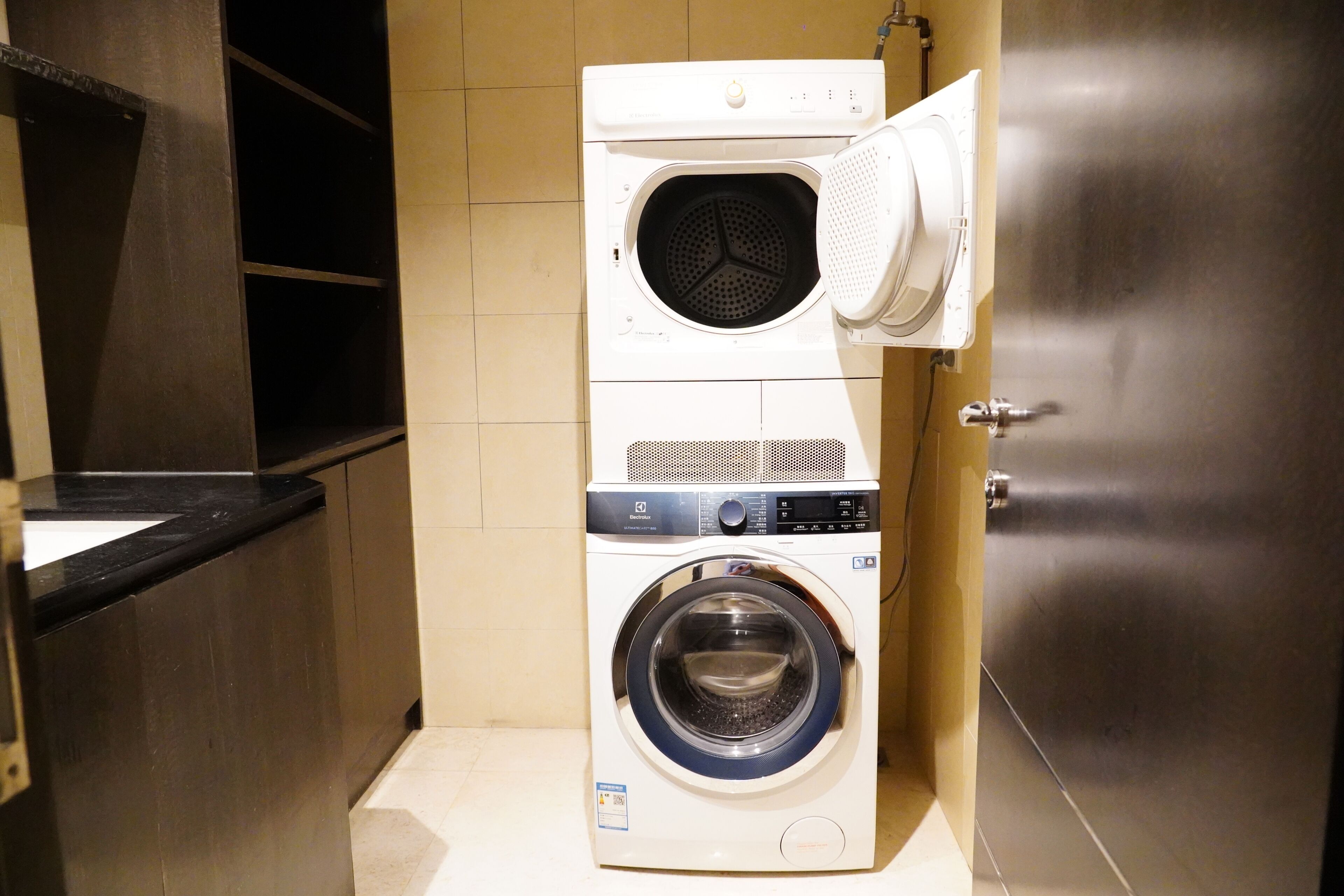 A compact, wall-mounted laundry machine with a front-loading washing compartment and a top-loading dryer, situated in a small, tiled laundry room with a wooden cabinet and a stainless steel door.