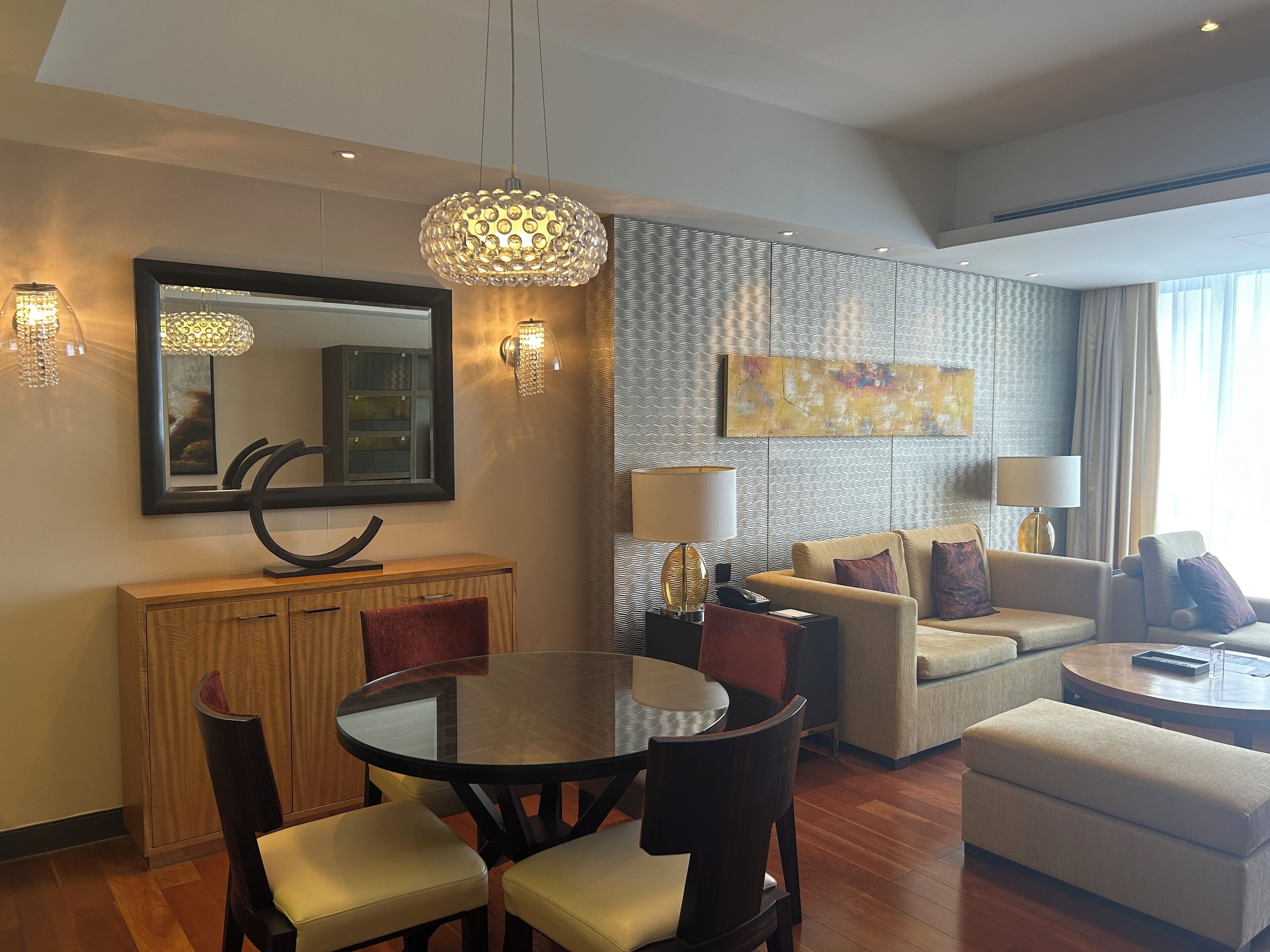 Quarto de hotel moderno com área de refeições, sala de estar com sofá e poltronas, mesa de jantar redonda e um espelho grande.