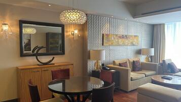 Quarto de hotel moderno com área de refeições, sala de estar com sofá e poltronas, mesa de jantar redonda e um espelho grande.