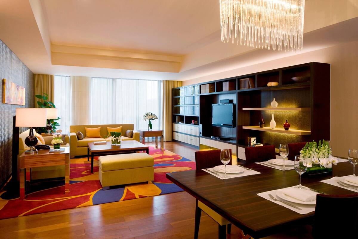 A modern hotel room with a living area, dining space, and a large bookshelf.