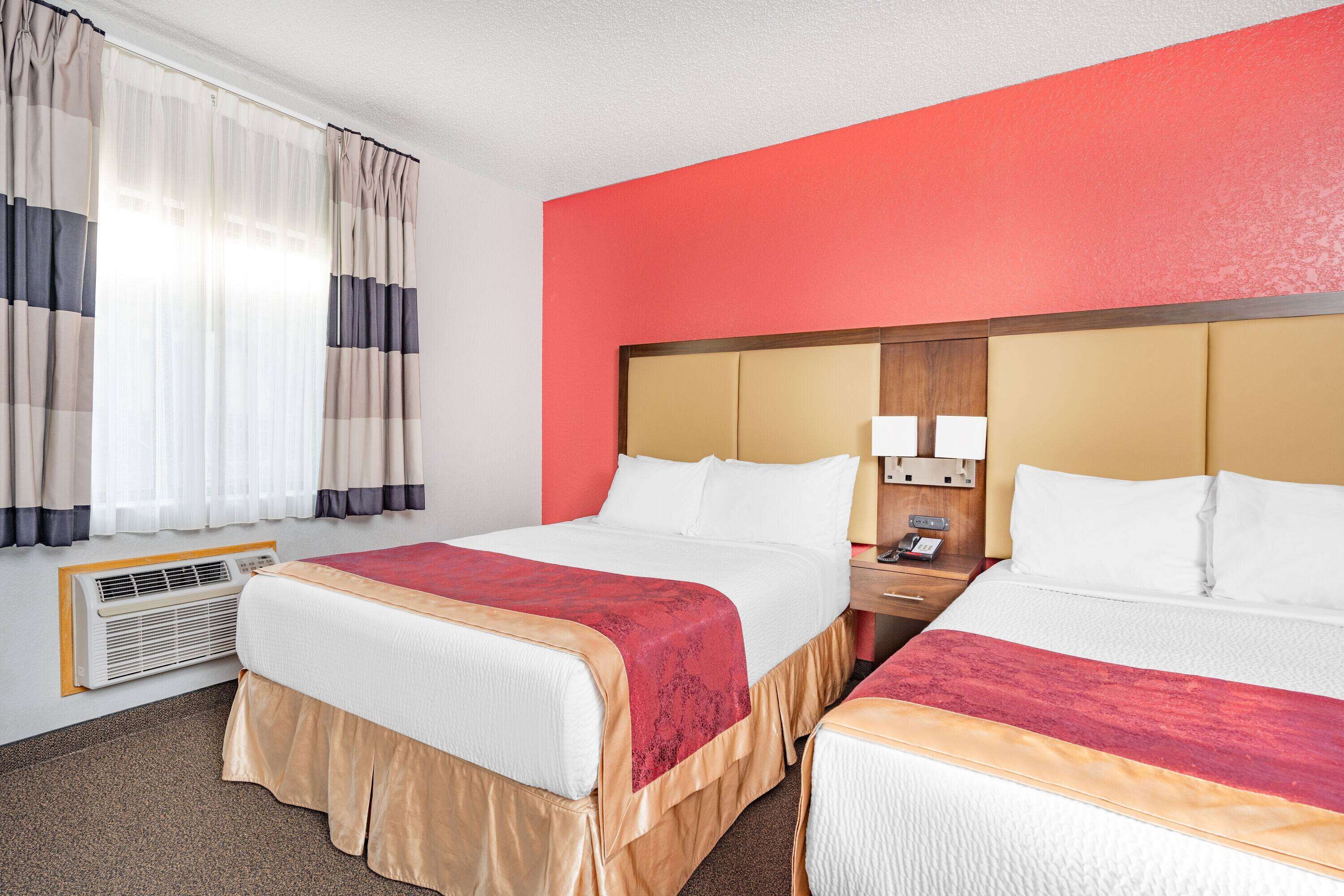 Une chambre d’hôtel avec deux lits, un mur d’accent rouge, une table de chevet avec une lampe et une fenêtre avec des rideaux.