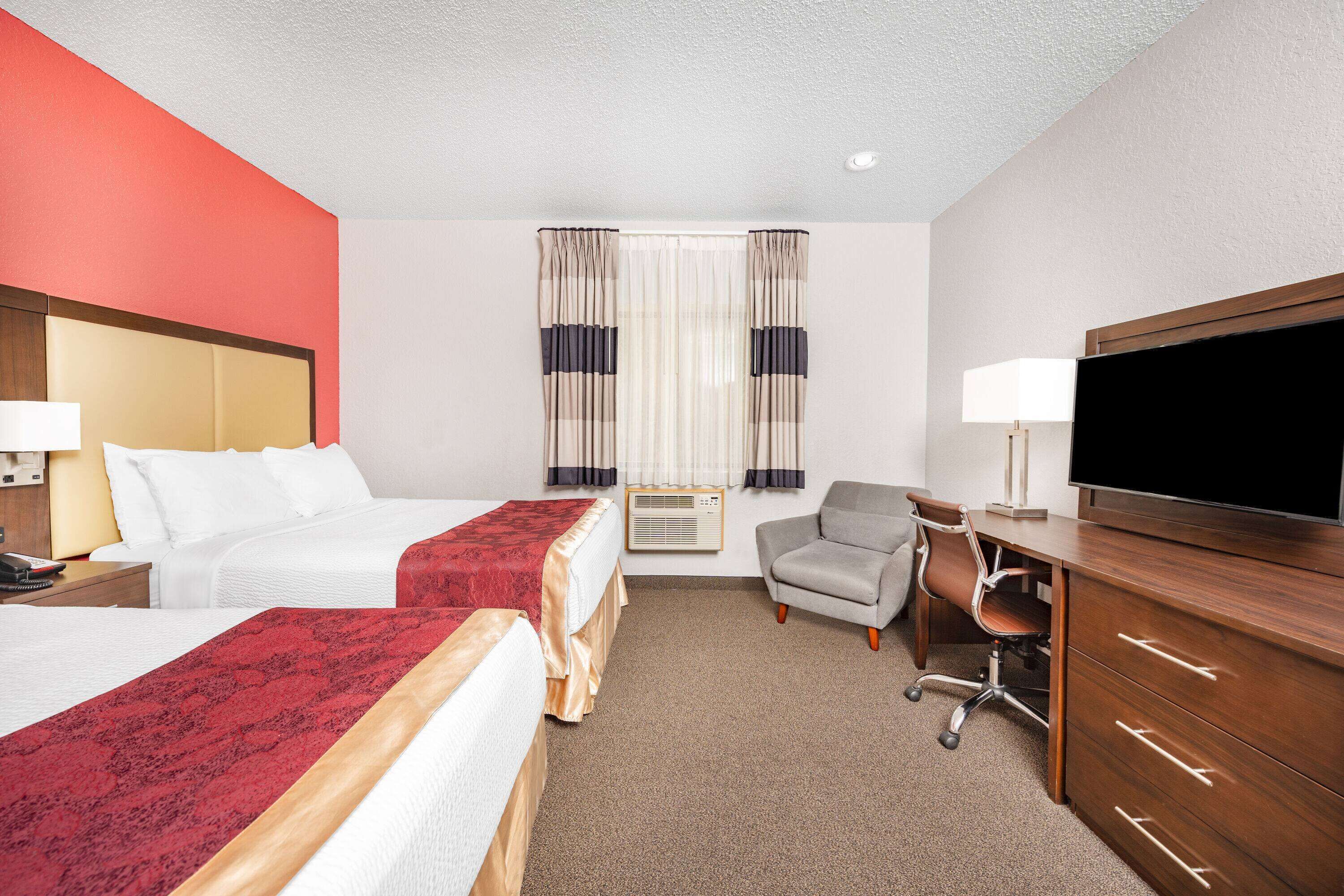 A hotel room with two beds, a desk, a chair, a TV, and an air conditioning unit.