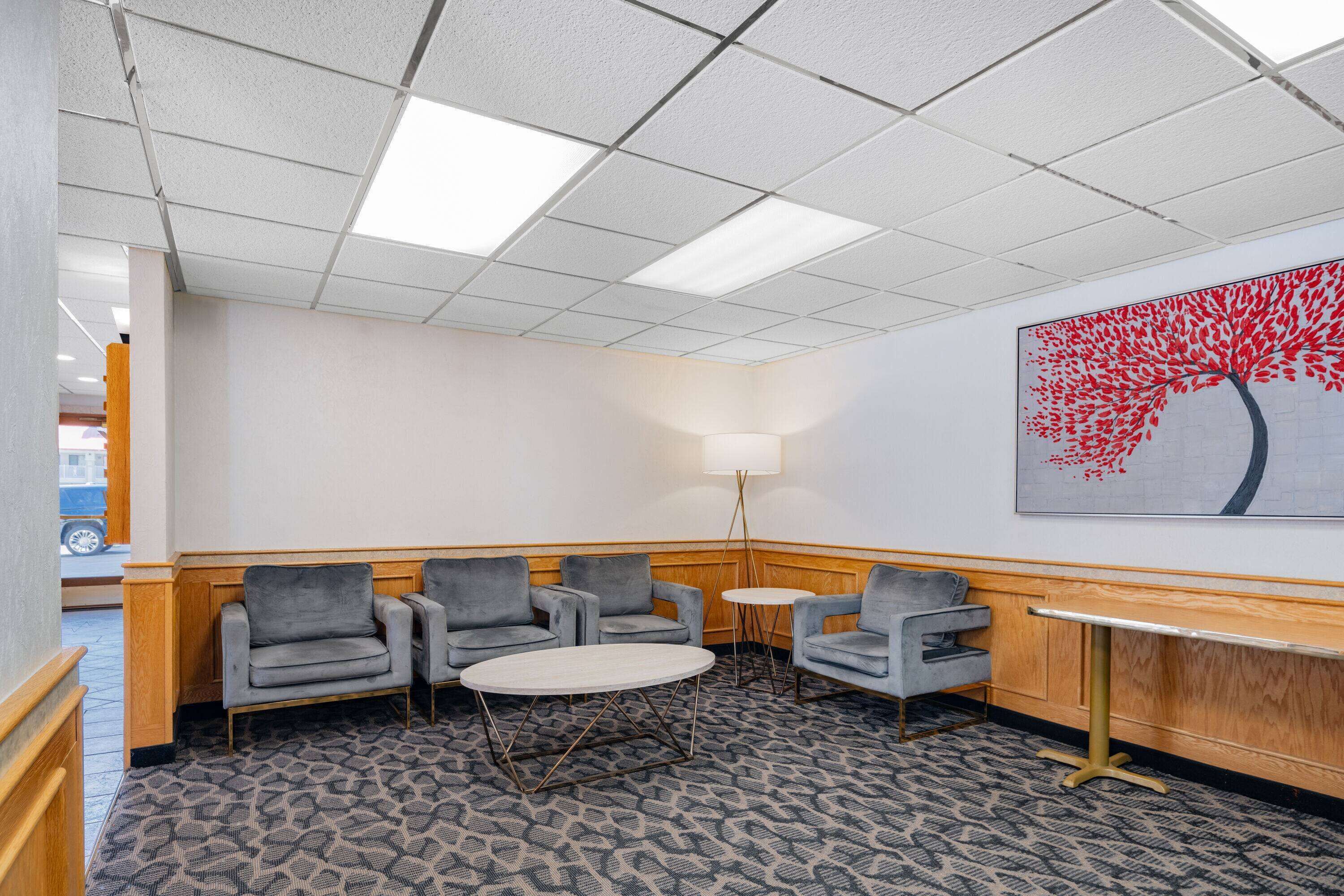 A hotel lobby with a patterned carpet, grey armchairs, a round table, a floor lamp, and a wall-mounted painting of a tree.