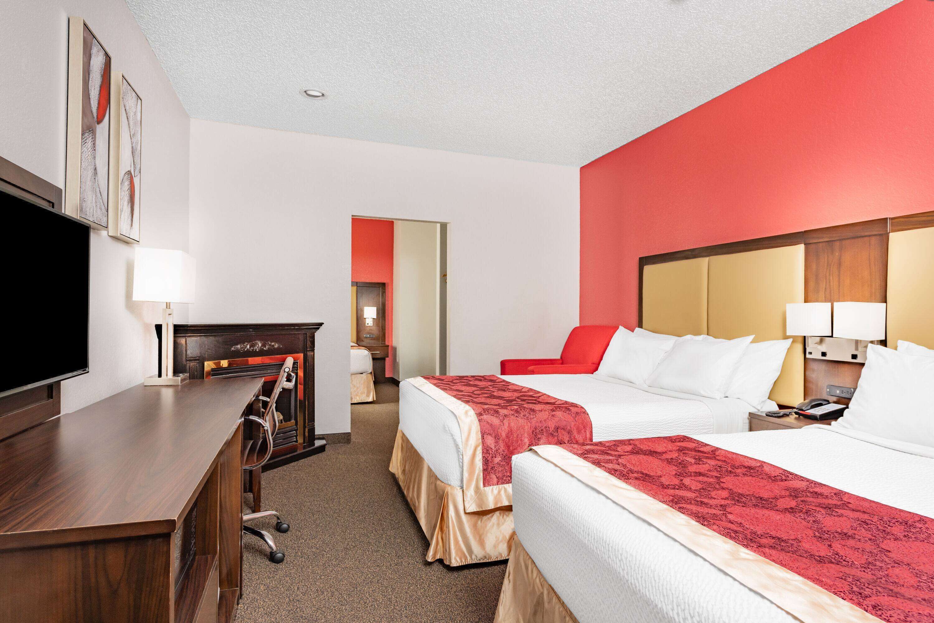 A hotel room with a wooden desk, two beds with red and white bedding, a TV, and a red accent wall.