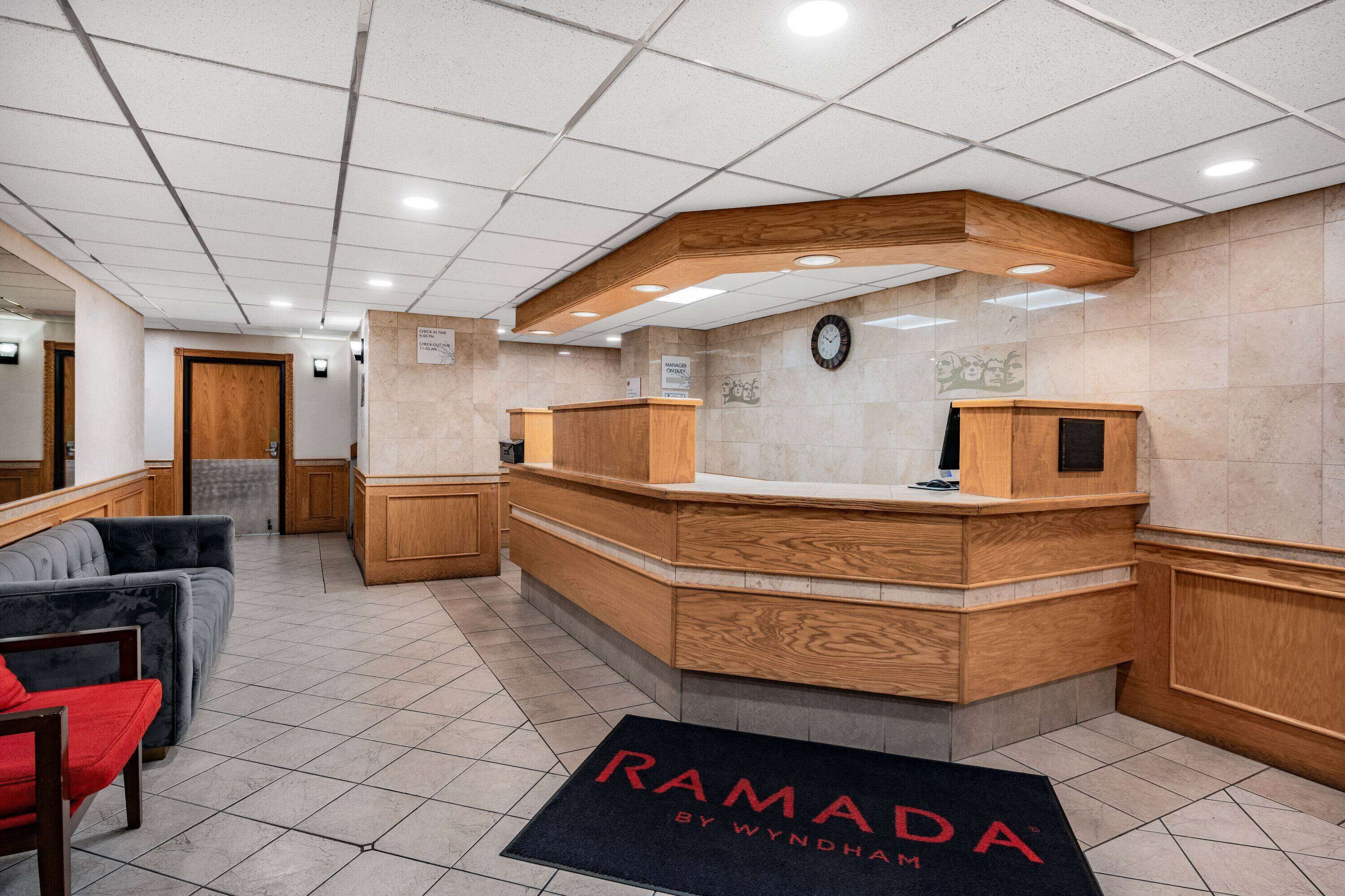 Un espace d’accueil avec un comptoir en bois, une horloge au mur et un paillasson sur lequel est écrit « RAMADA ».