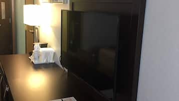 A hotel room with a dark wooden desk, a flat-screen TV, a lamp, and a small table with a remote control and a menu.