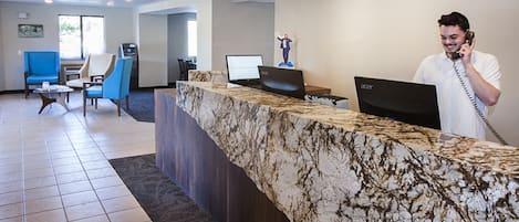 A hotel reception area with a marble counter, two laptops, a telephone, and seating with blue and beige chairs.
