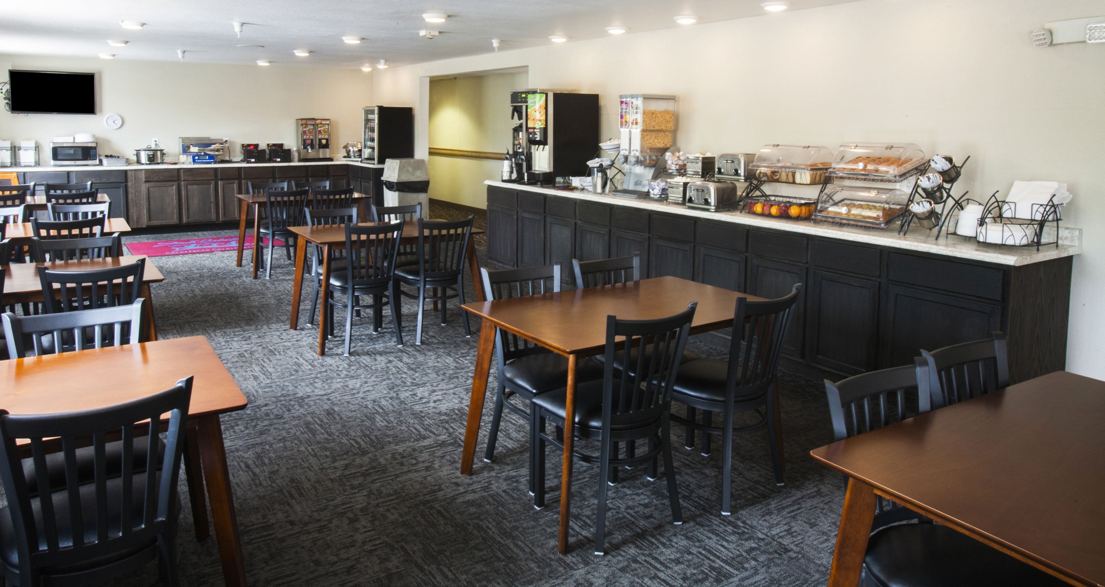 A hotel dining area with multiple tables and chairs, a kitchen counter with various appliances, and a buffet setup.