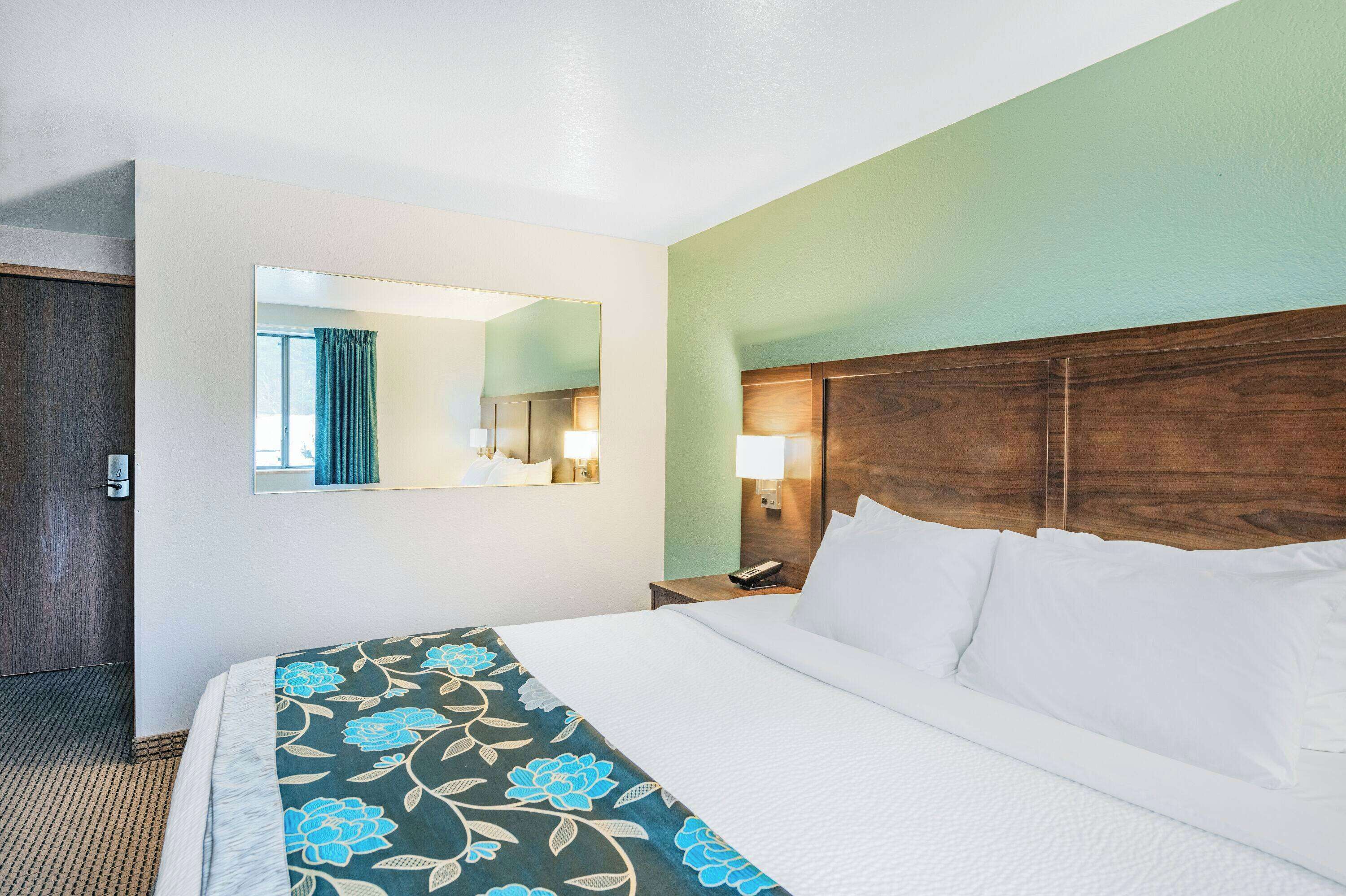 Een hotelkamer met een groot bed, een houten hoofdeinde, een nachtkastje met een lamp, een spiegel aan de muur en een deur links.