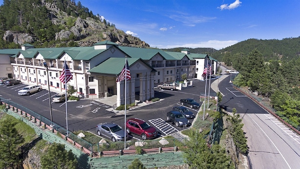 A multi-story hotel with a green roof, parking lot, and American flags. - Baymont by Wyndham Keystone Near Mt. Rushmore (Keystone)