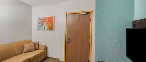 A room with a wooden door, a beige sofa with pillows, a painting on the wall, and a television on a stand.